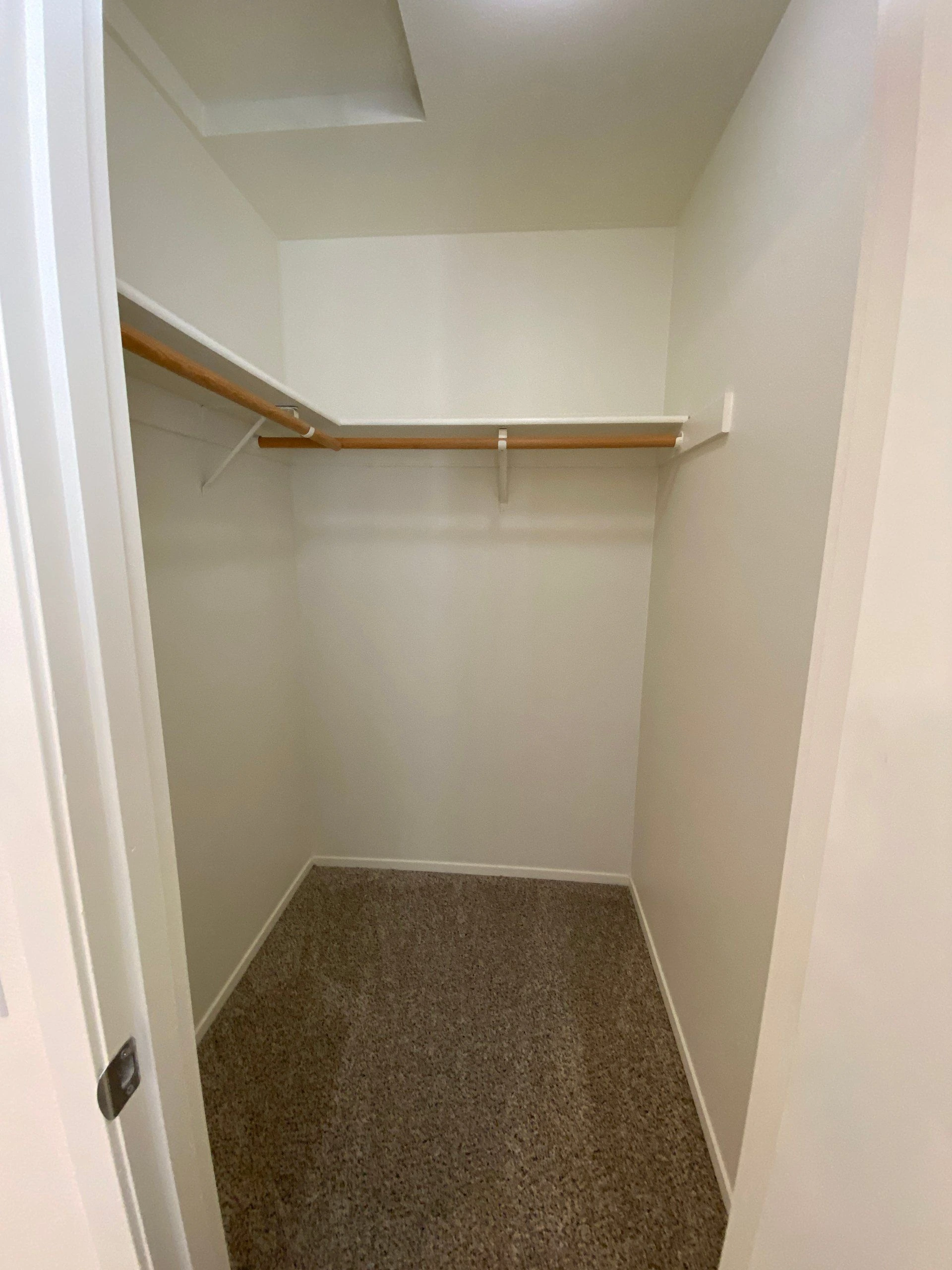 An empty carpeted closet with two wooden hanging rods beneath a white shelf and sloped ceiling.