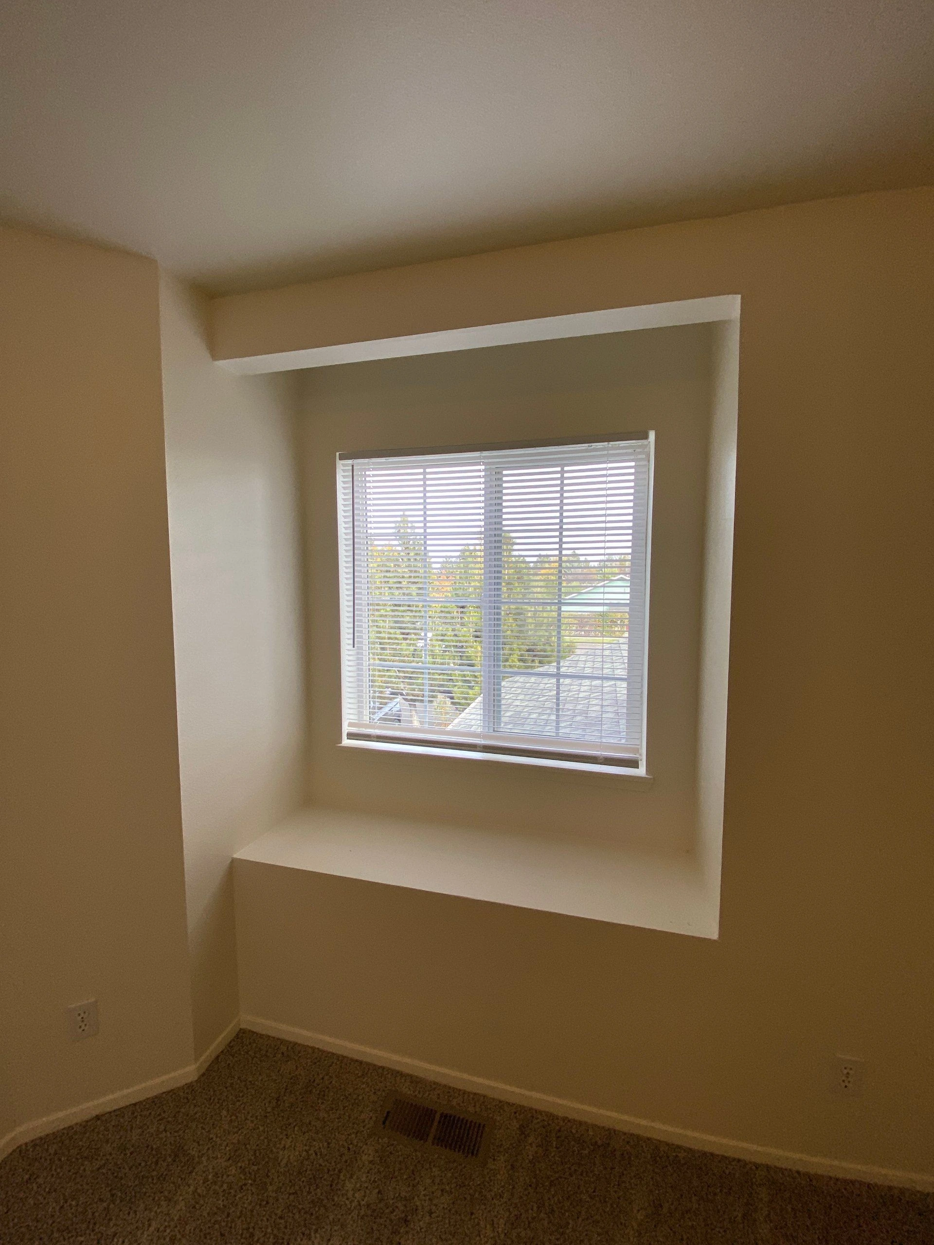 A recessed window with white blinds sits above a carpeted floor between beige walls.