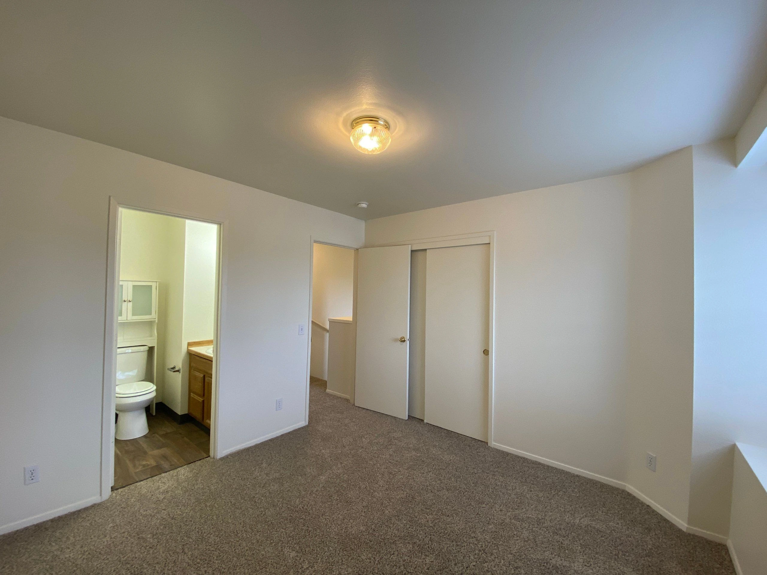An empty carpeted room has a lit ceiling fixture, an open bathroom doorway with a toilet and vanity, and sliding closet doors.