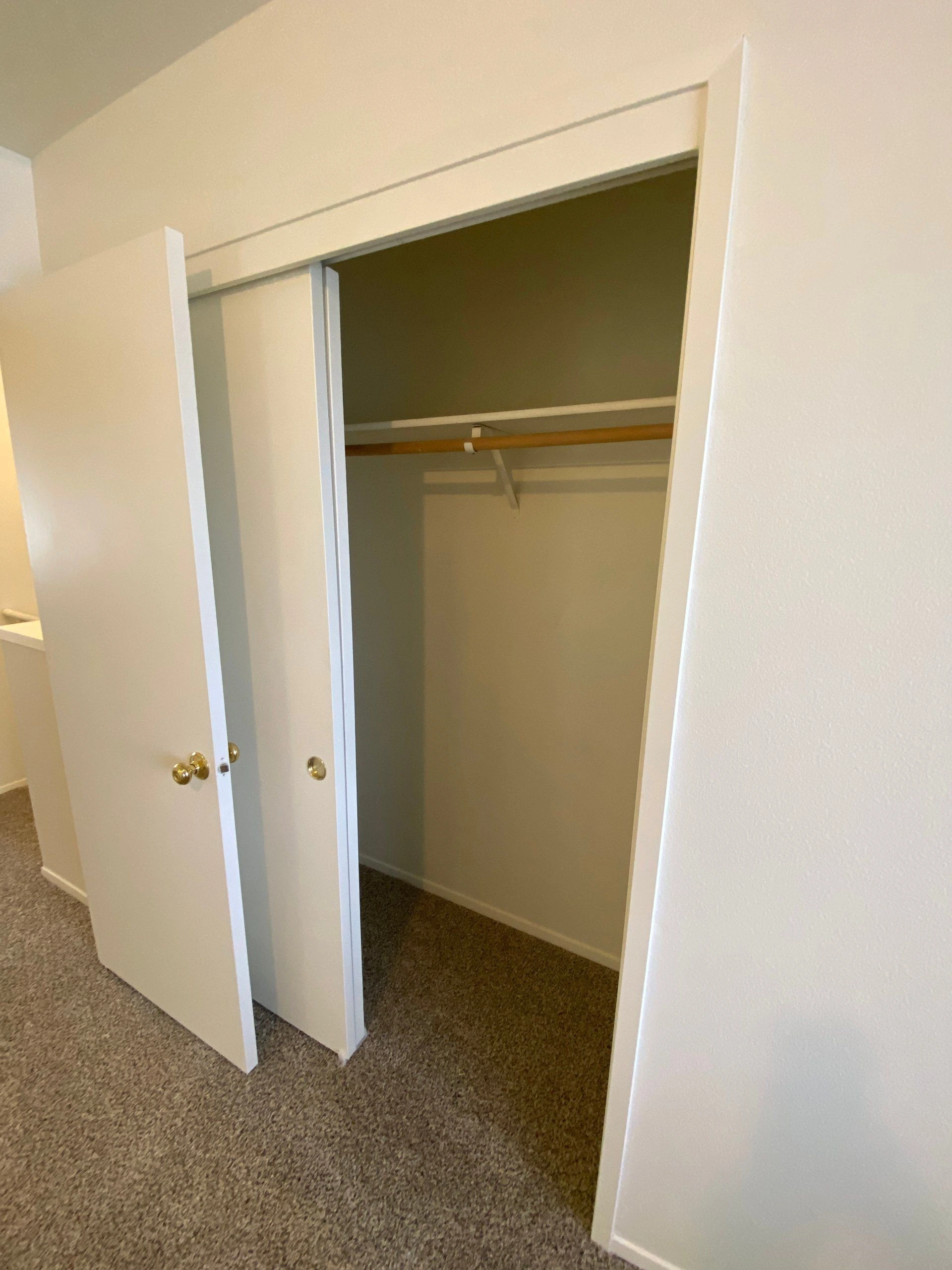An open closet has white sliding doors, a wooden hanging rod, an upper shelf, and carpeted flooring.