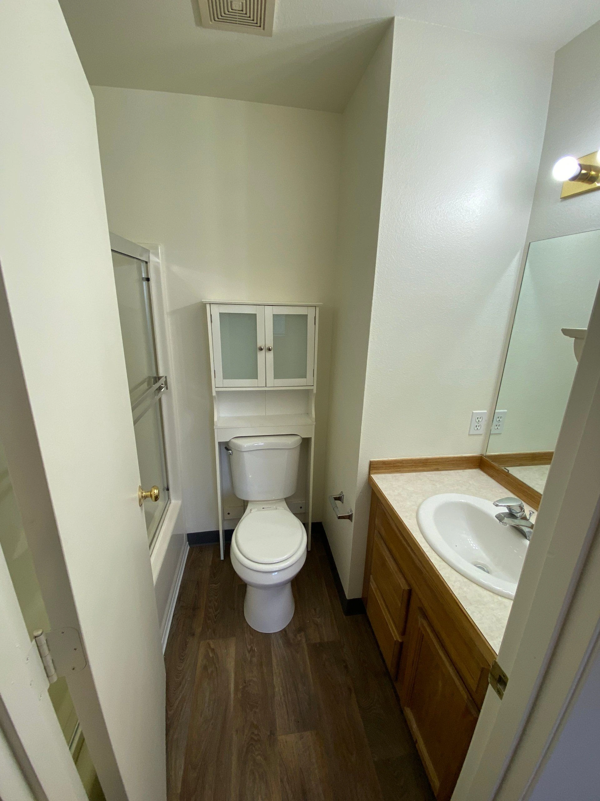 A narrow bathroom has a toilet beneath a wall cabinet, a sink in a wood vanity, and a shower enclosure.