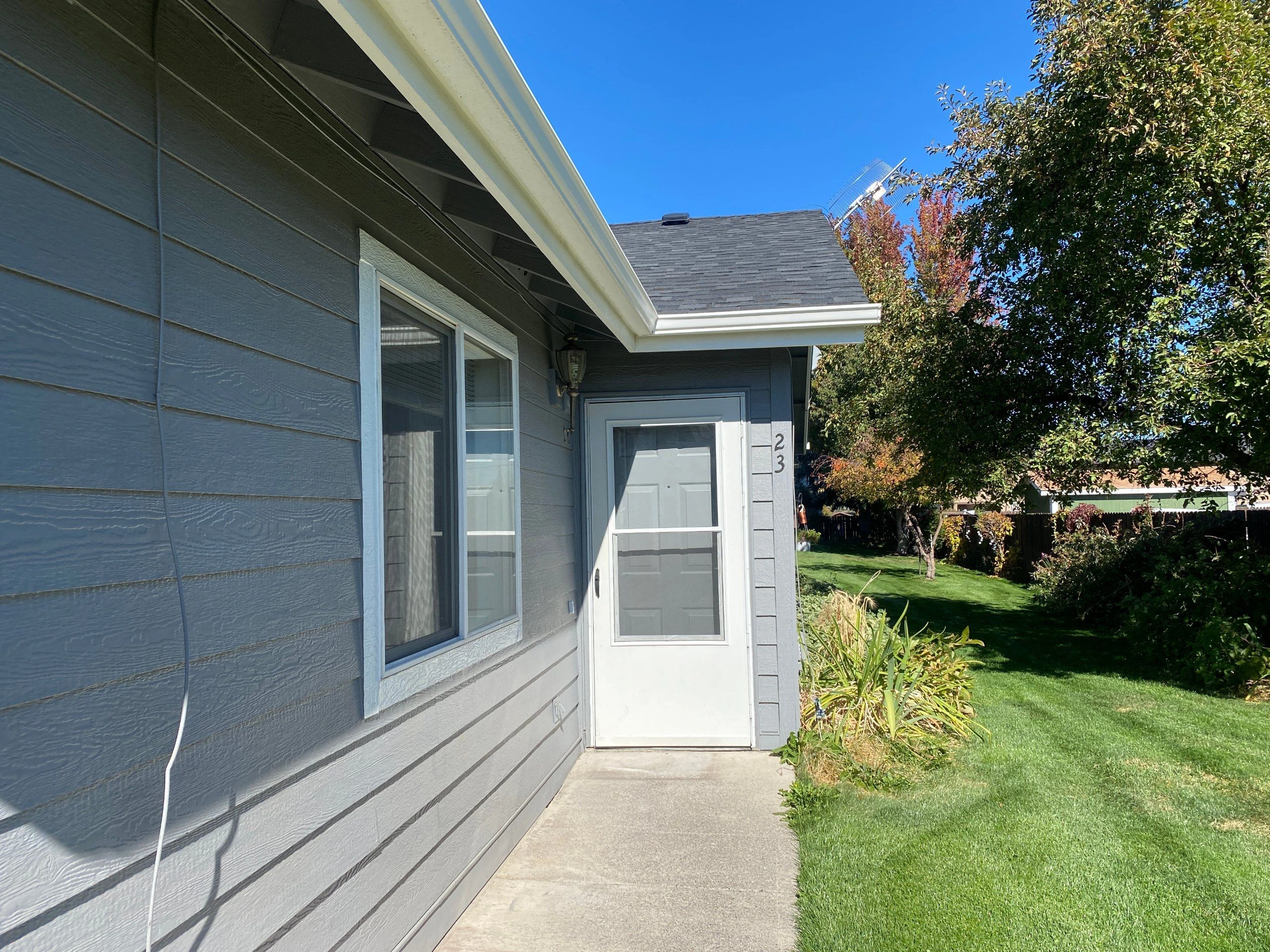 A white screen door and window on a gray-sided house open onto a concrete path beside a green lawn and trees.