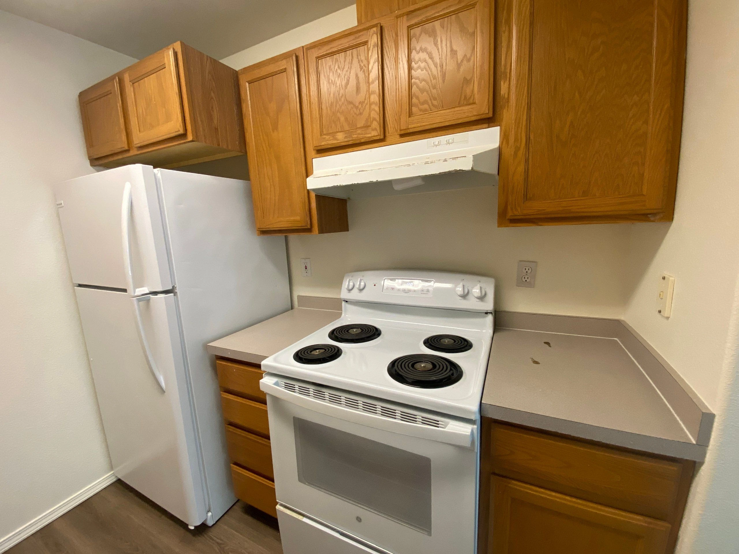 A white refrigerator and four-coil electric stove sit beneath wooden cabinets, beside beige countertops and a range hood.