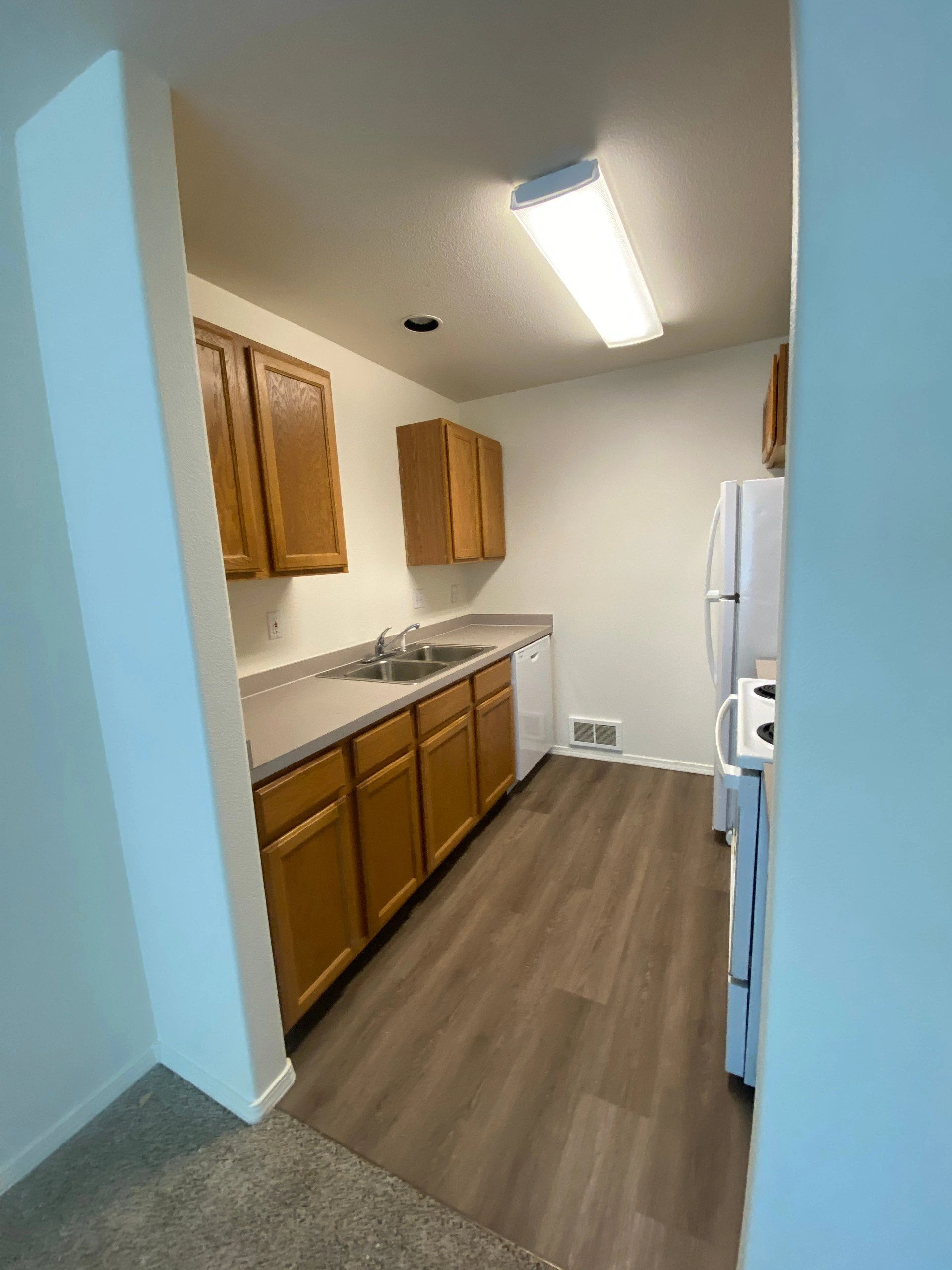 A narrow kitchen has wooden cabinets, a double sink, a long counter, and white appliances.