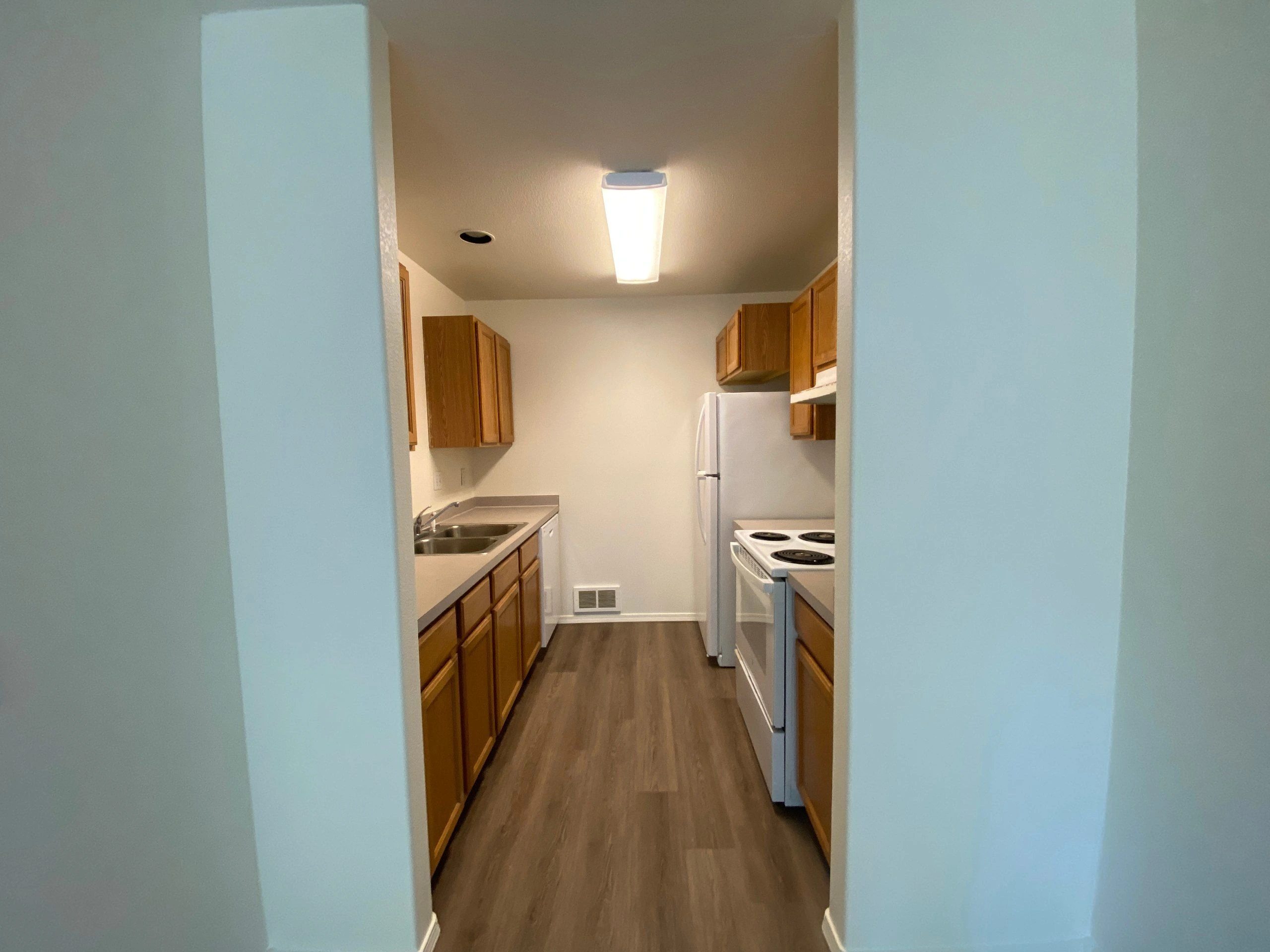 A narrow kitchen has wooden cabinets, a double sink, a refrigerator, and an electric stove.