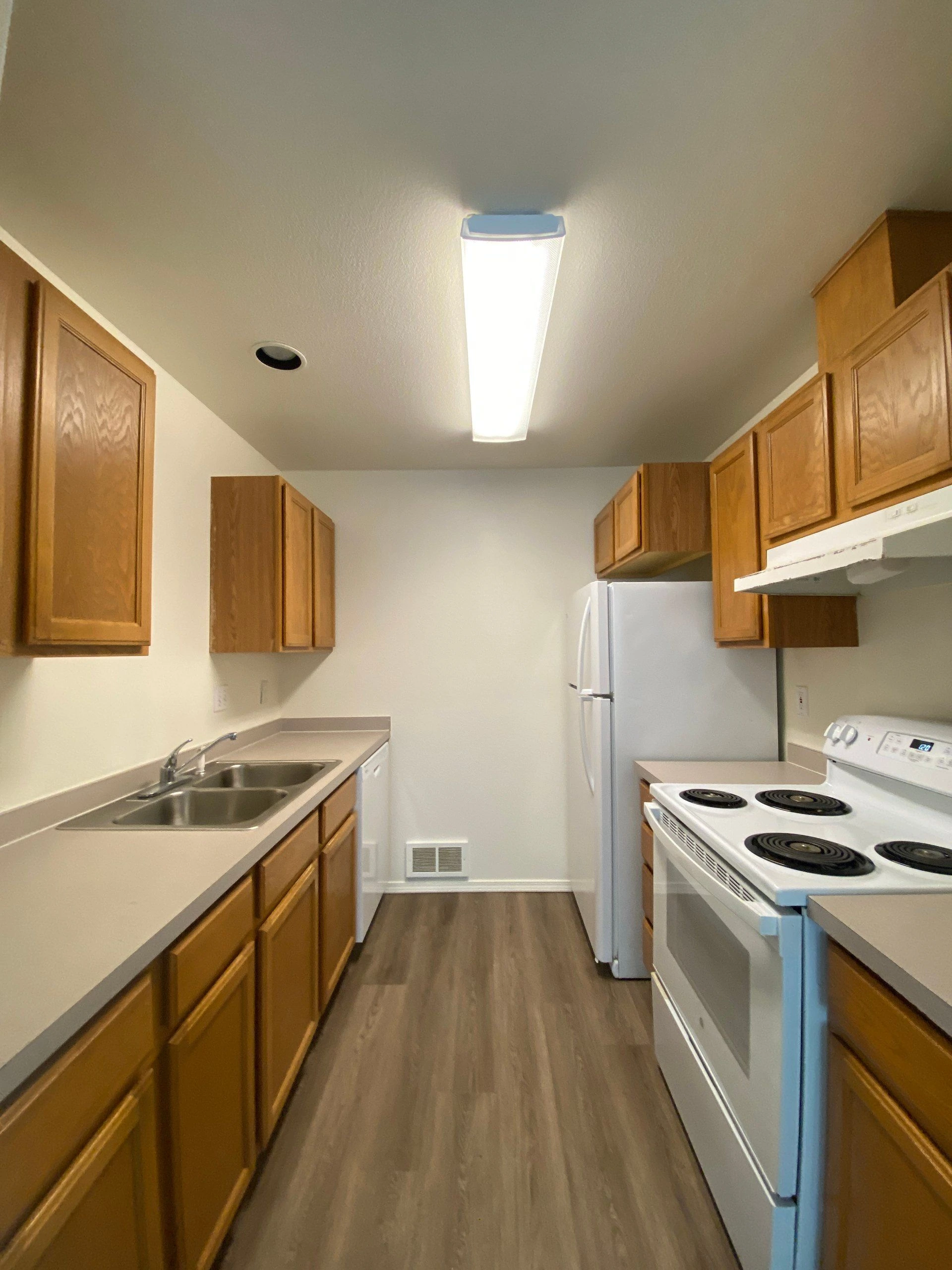 A narrow kitchen with wood cabinets, a double-basin sink, a refrigerator, and an electric stove.