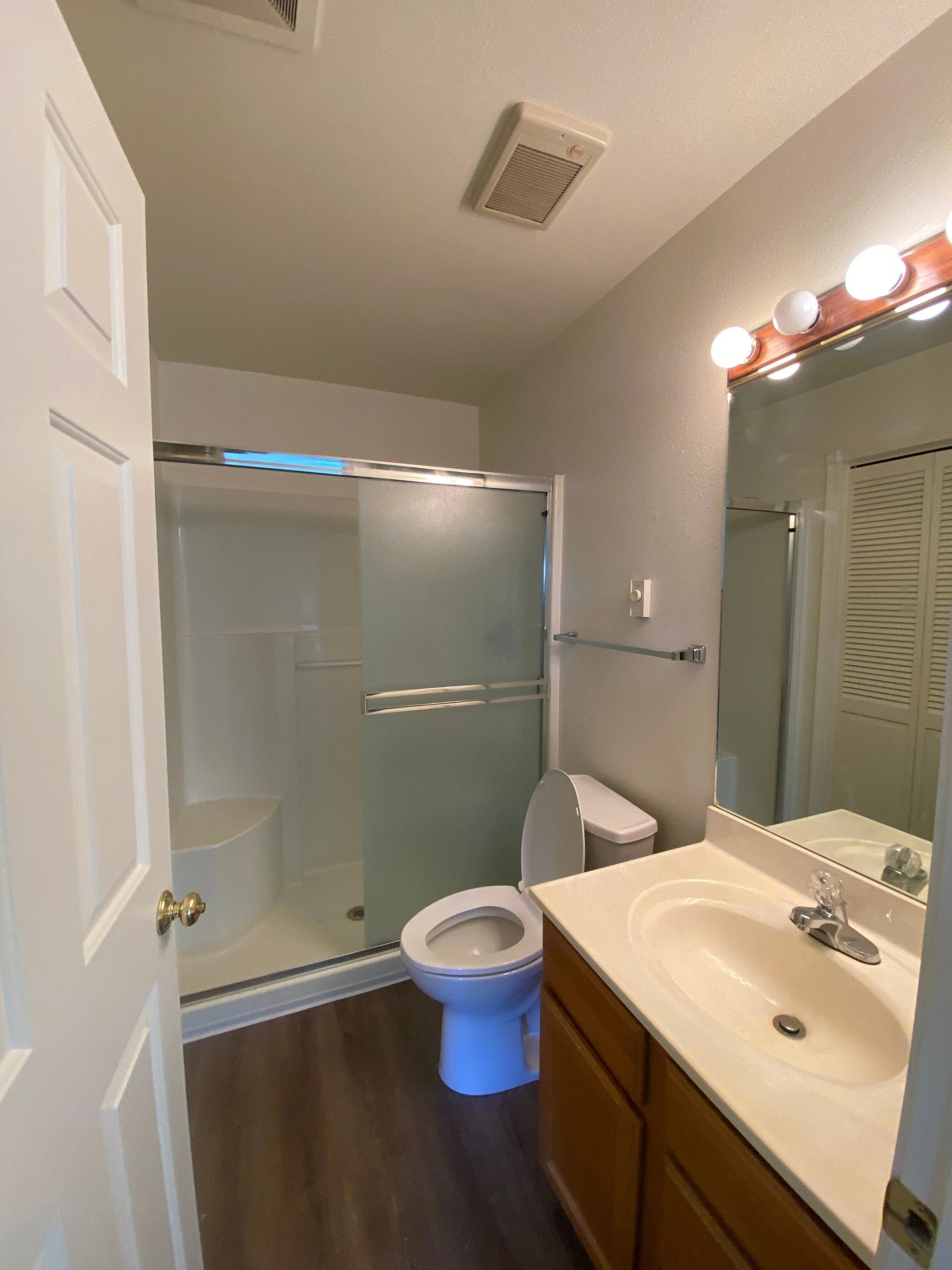 A bathroom with a frosted-glass shower door, toilet, sink vanity, and a large mirror with lights.