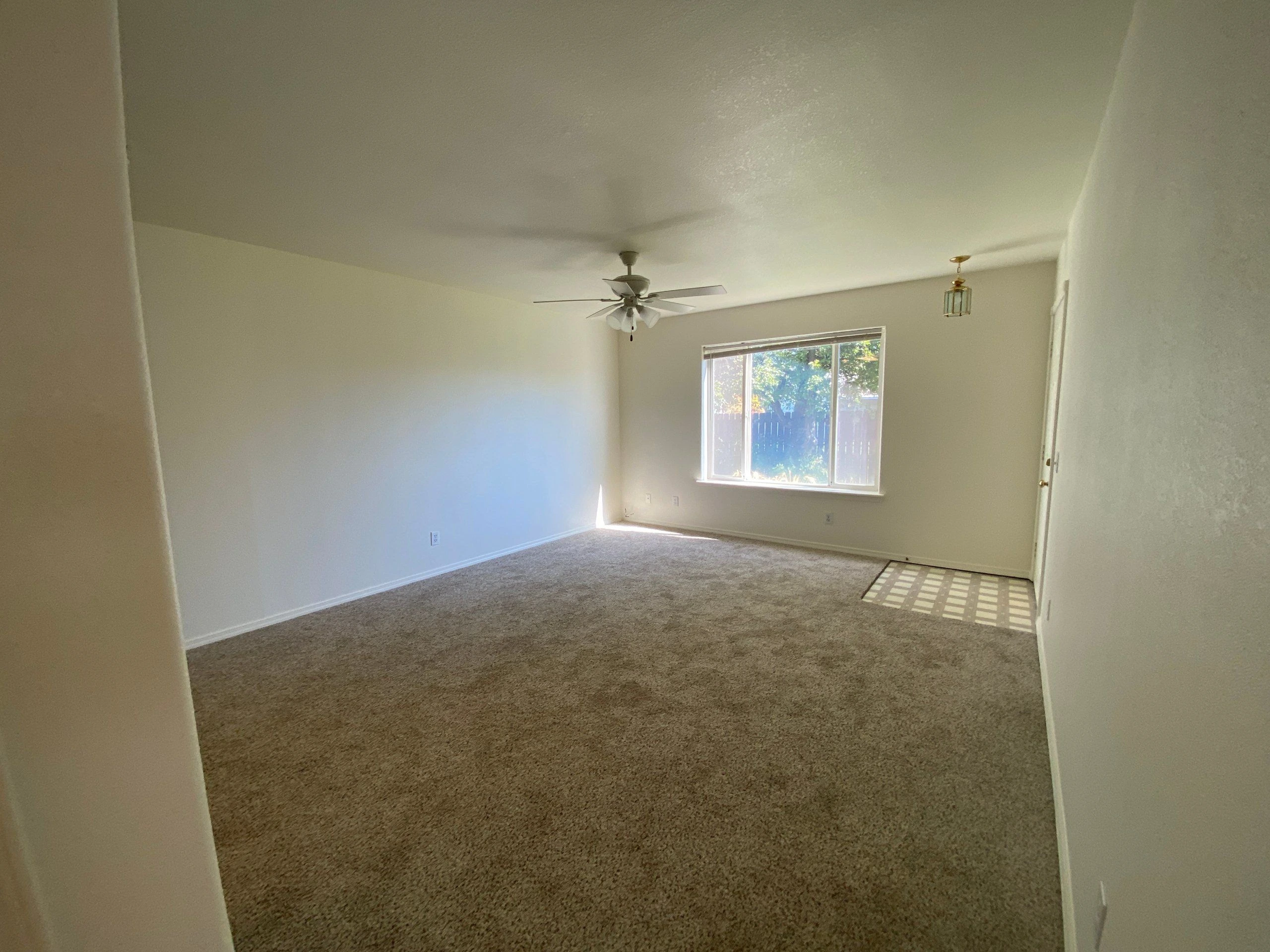 An empty carpeted room has pale walls, a ceiling fan, a hanging light, and a window with blinds.