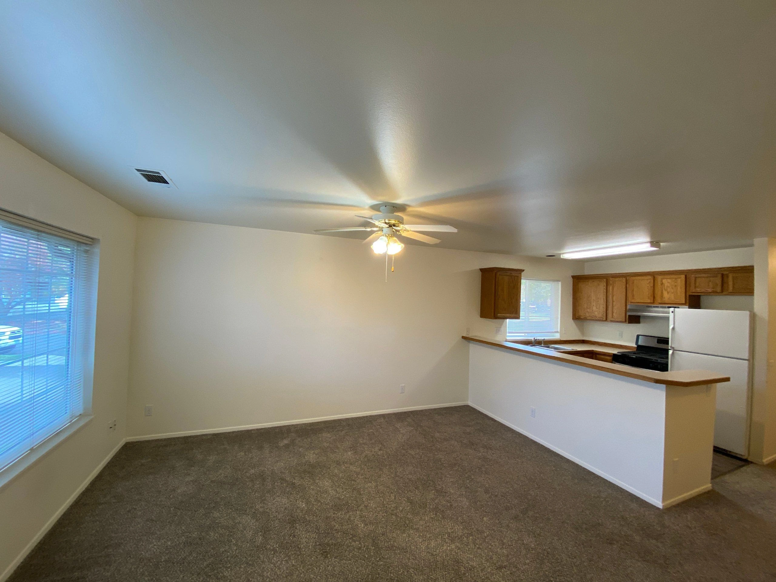 Carpeted living area opening to a kitchen with wood cabinets, appliances, a counter, and a ceiling fan.