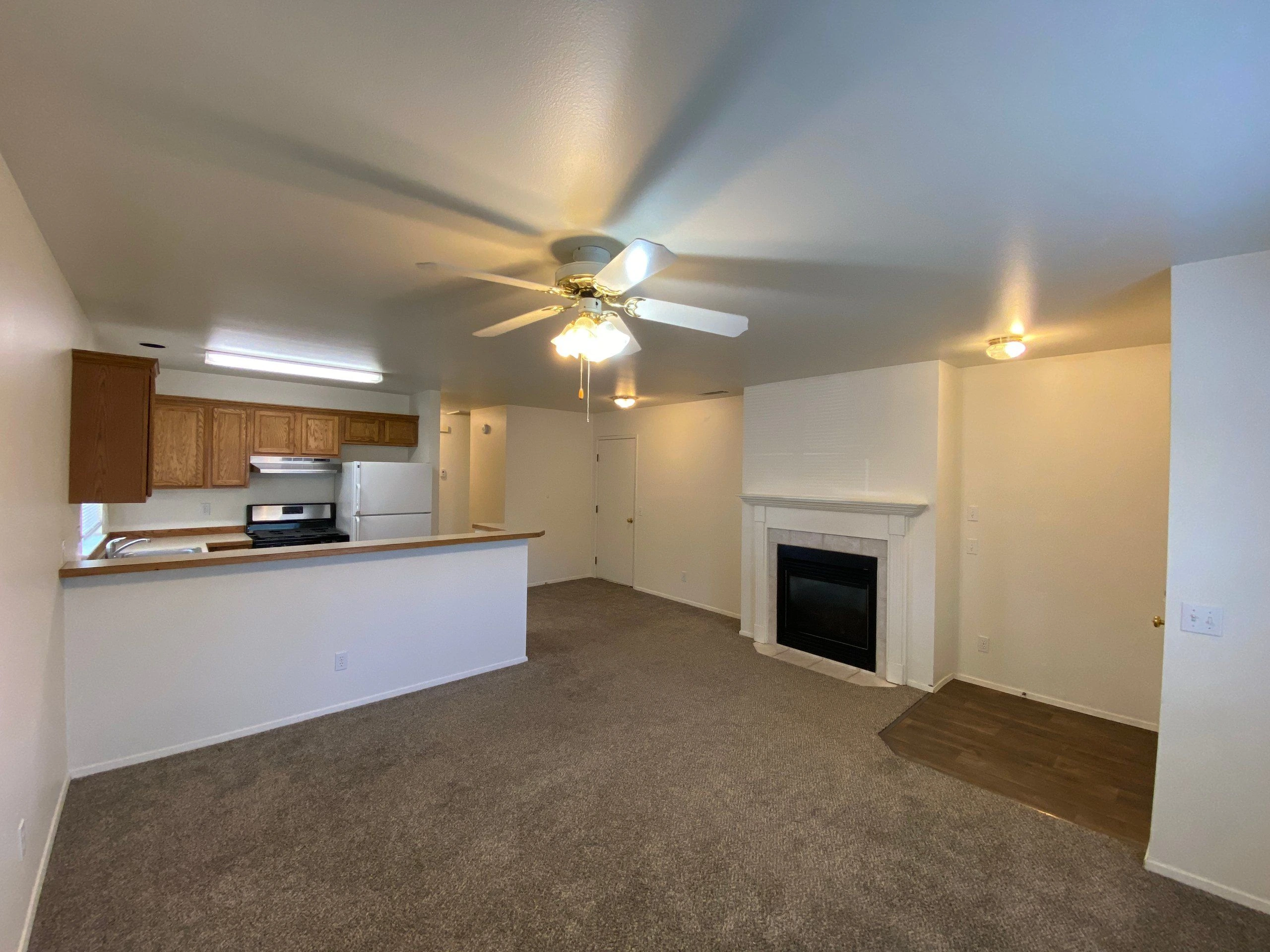 An open room has a kitchen counter with wood cabinets and appliances, a ceiling fan, a fireplace, and carpet beside a wood floor section.