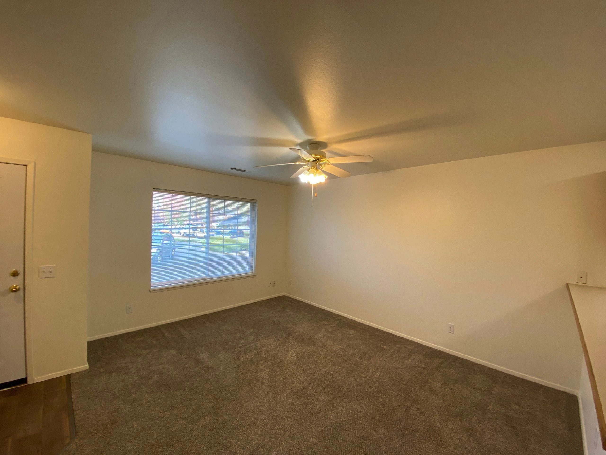 An empty room with carpet, a window with blinds, a ceiling fan with a light, and a door.