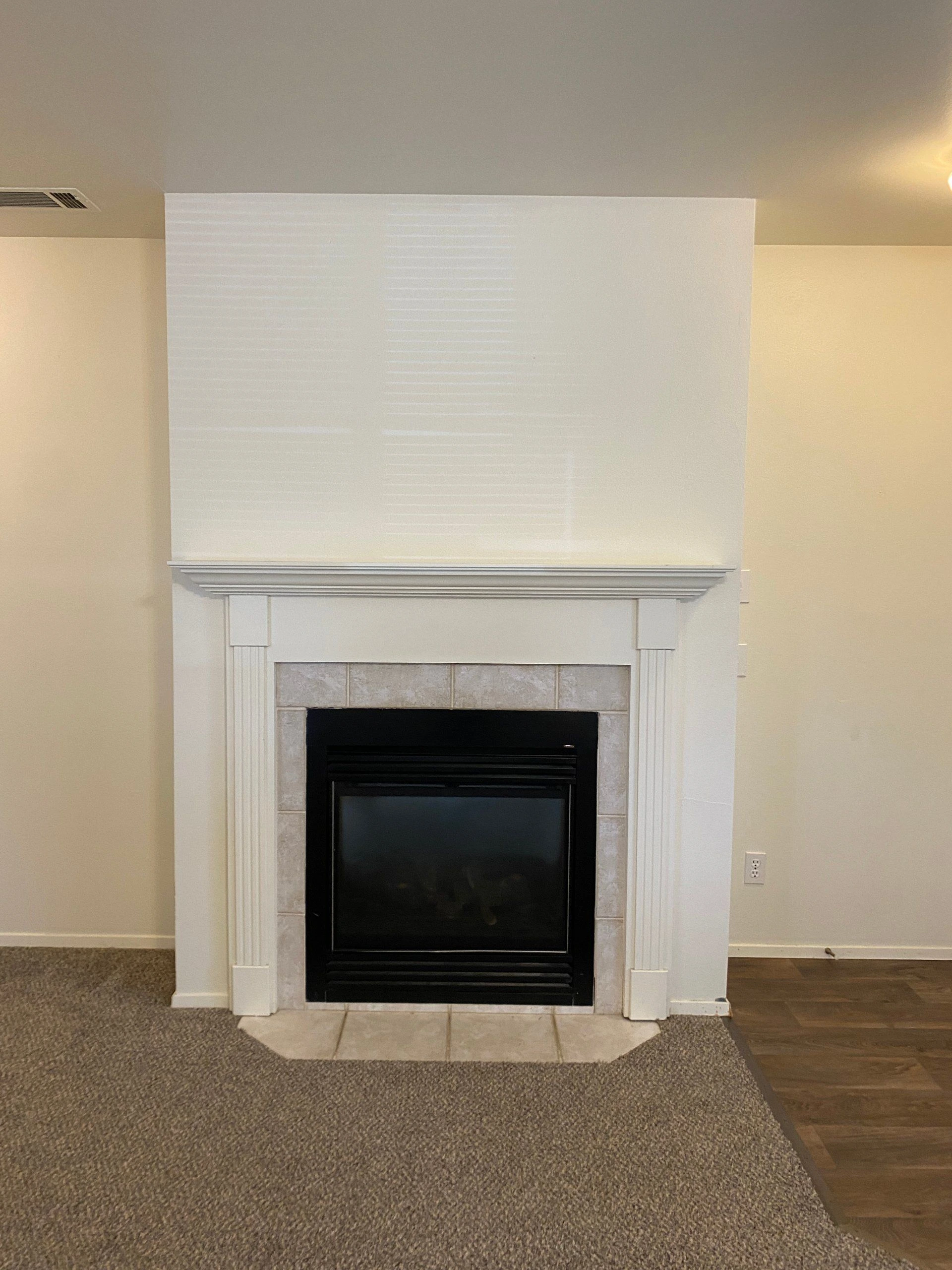 A white mantel surrounds a black fireplace insert with light tile; carpet covers the foreground beside wood flooring.
