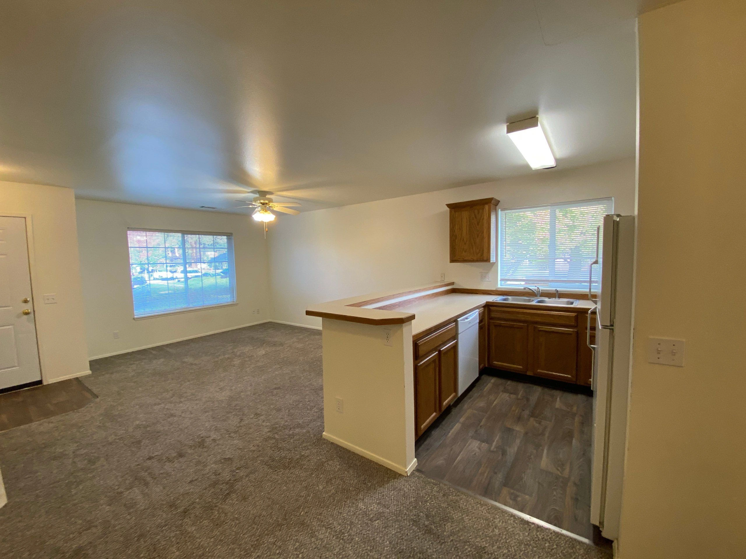 An open carpeted room connects to a kitchen with dark wood cabinets, a peninsula counter, sink, dishwasher, and refrigerator.