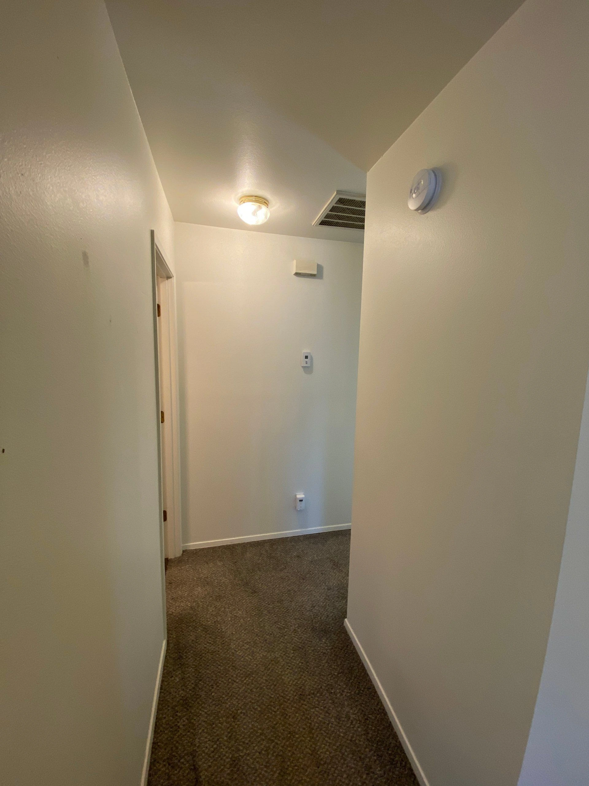 A narrow hallway with brown carpet, pale walls, a ceiling light, an air vent, and an open doorway.