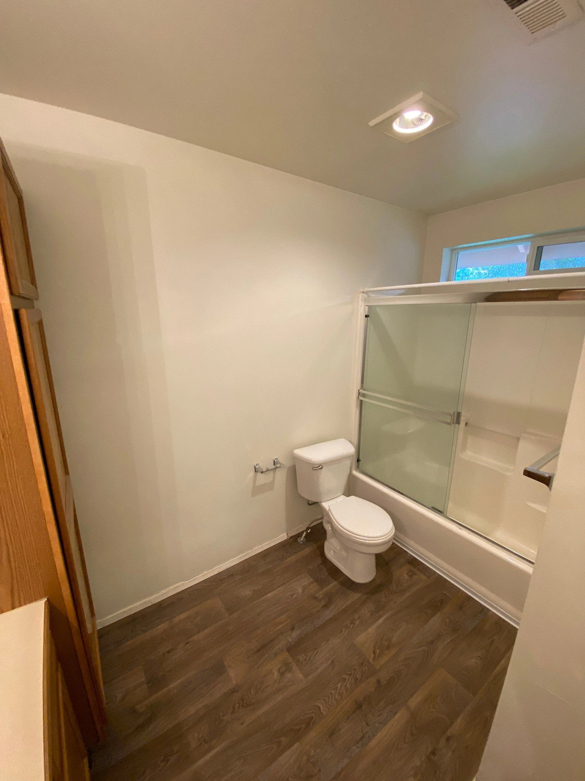 A bathroom with a toilet beside a bathtub enclosed by sliding glass doors, a small window, and dark wood-patterned flooring.