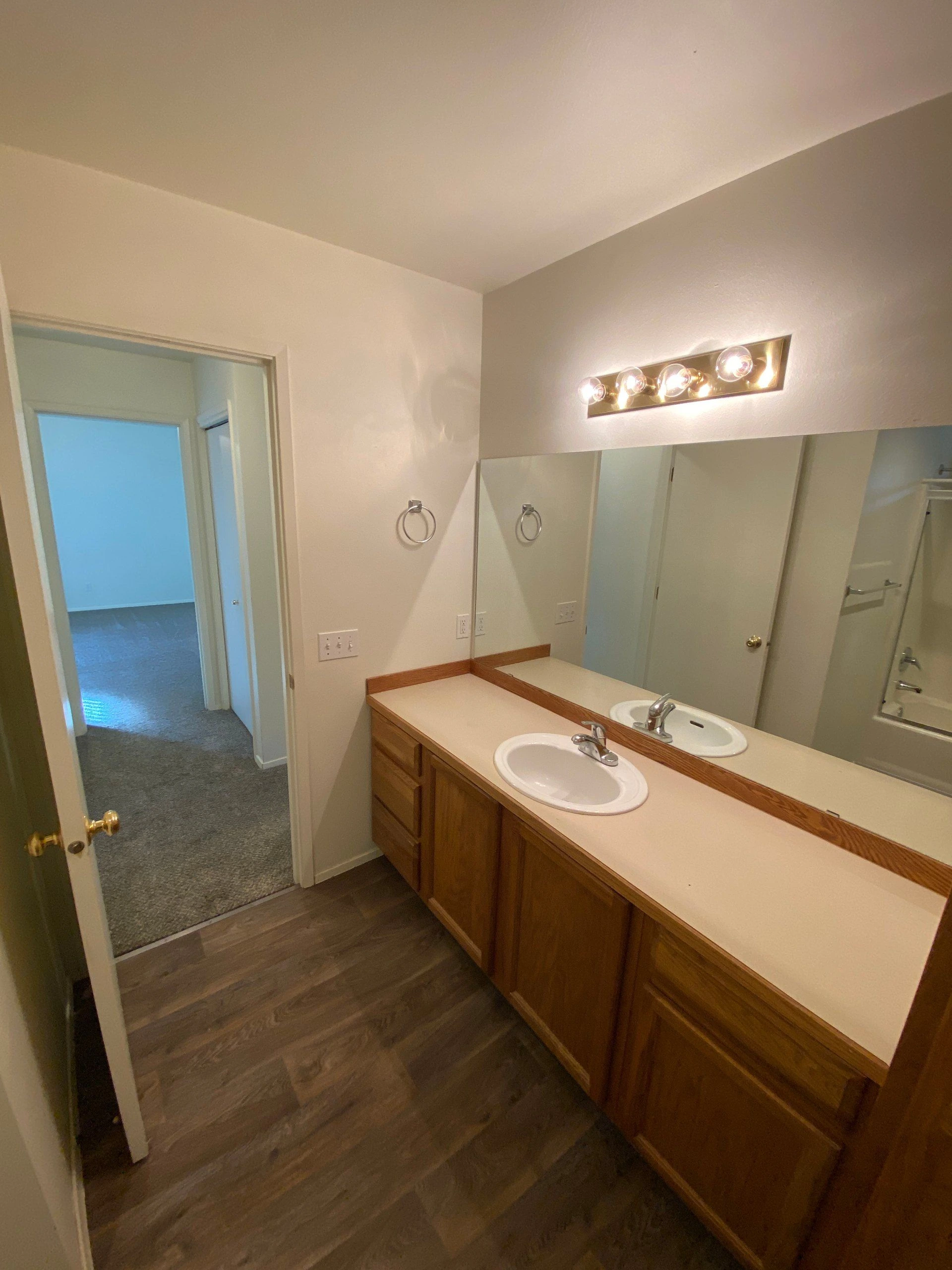 A bathroom vanity has two sinks, wooden cabinets, a long mirror, and a lit four-bulb fixture above it.