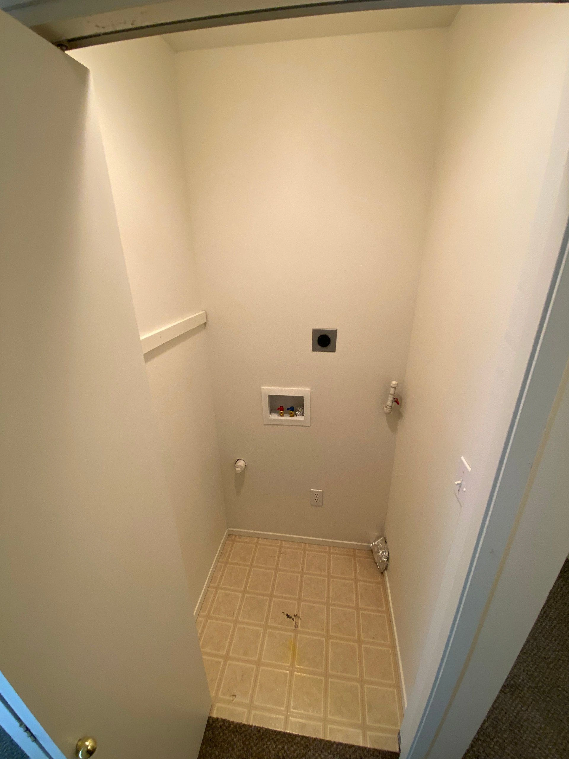 A narrow empty alcove has a square-patterned floor, wall-mounted pipes and valves, an electrical outlet, and a short shelf.