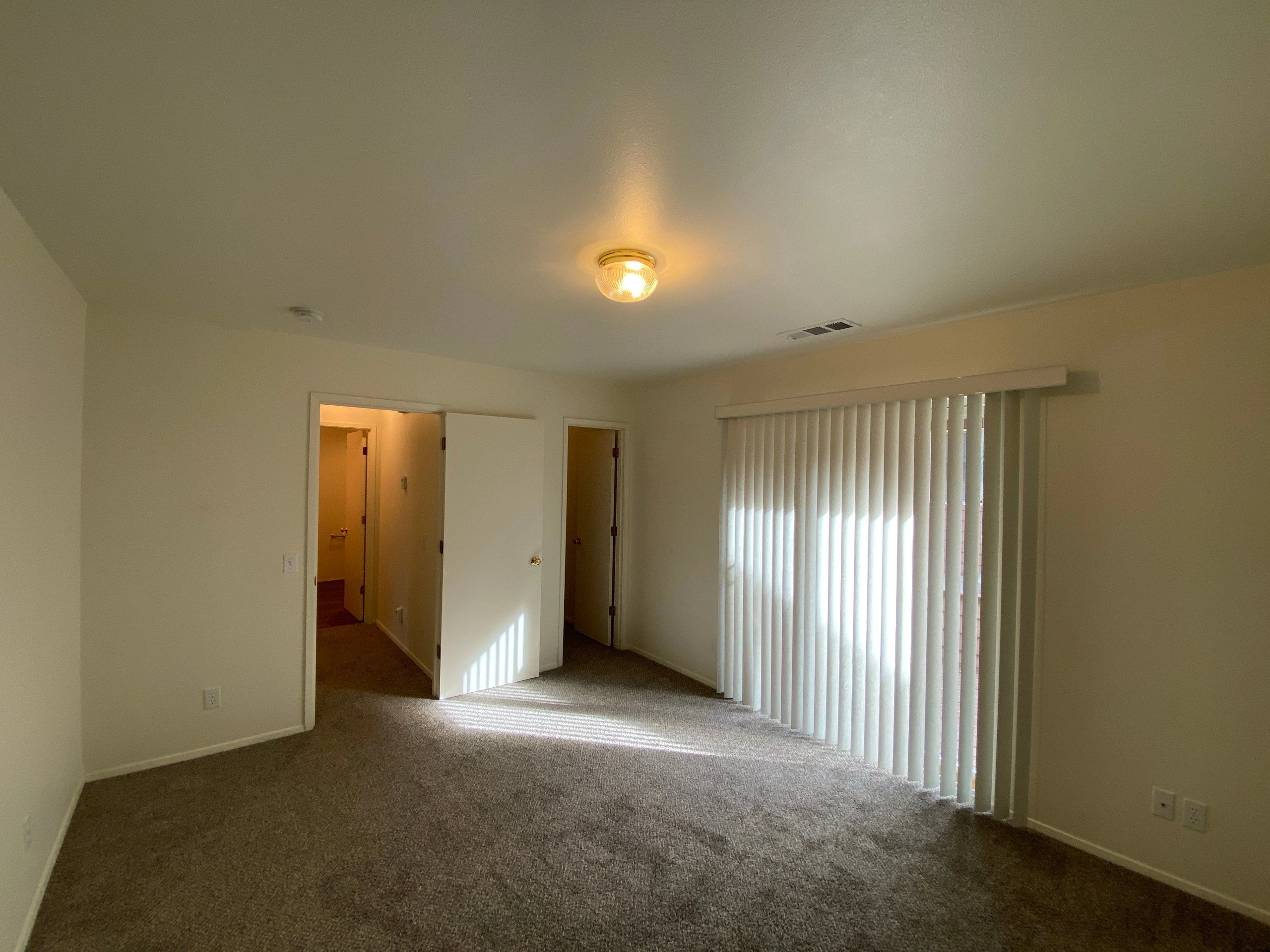An empty carpeted room has open doors, vertical blinds over a window, and a lit ceiling fixture.