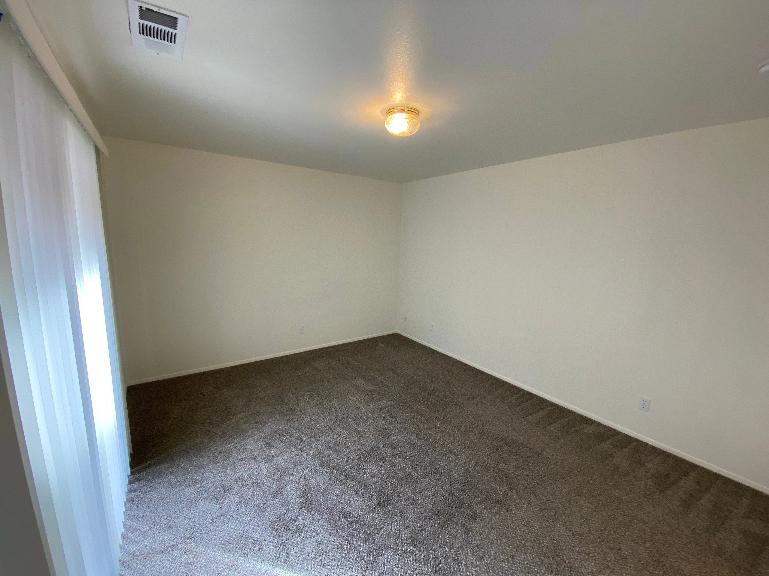 An empty room has beige walls, gray carpet, vertical blinds, and a lit ceiling fixture.