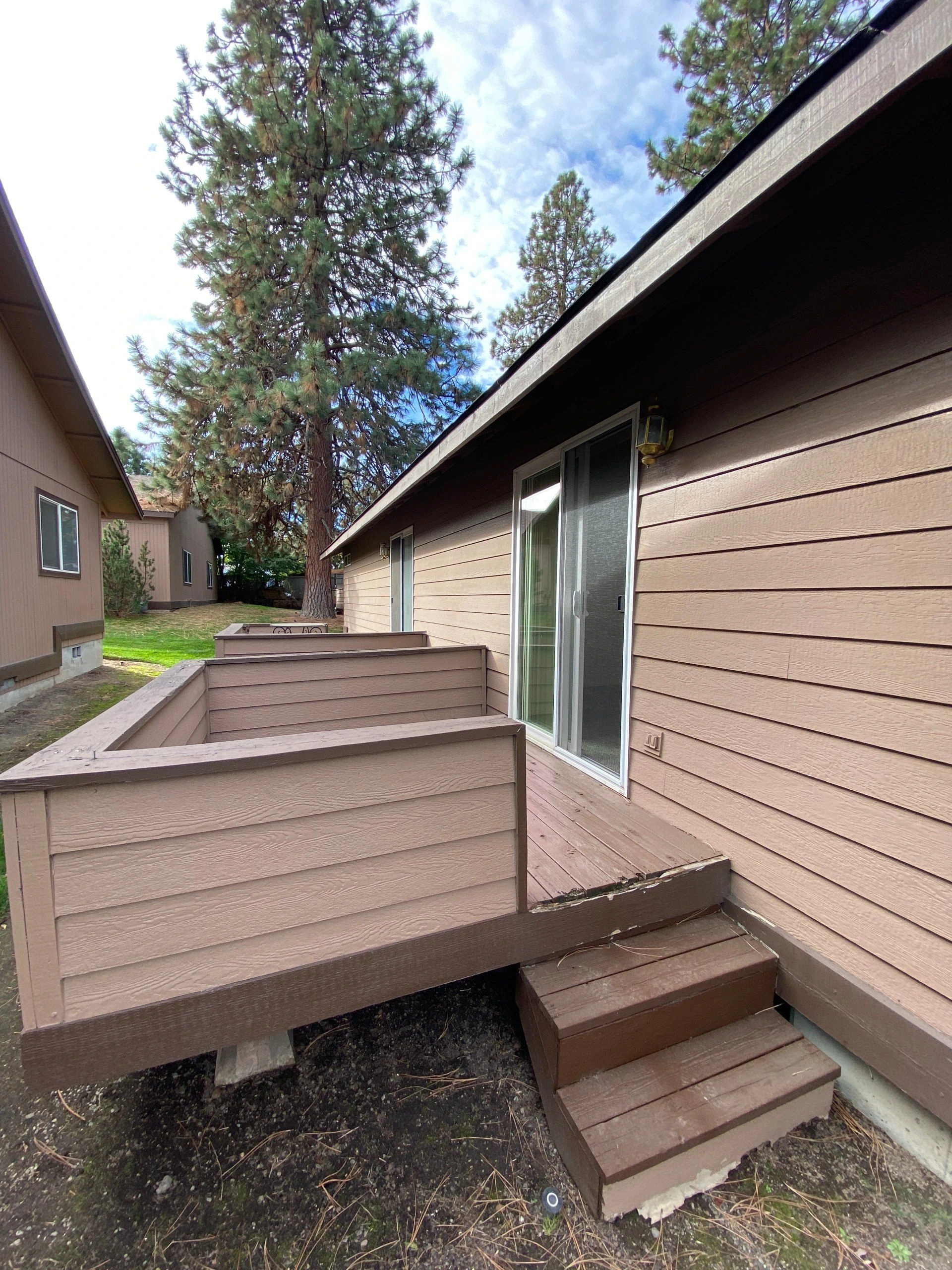 A wooden deck with railings and steps runs beside a tan-sided building with a sliding glass door, beneath tall trees.