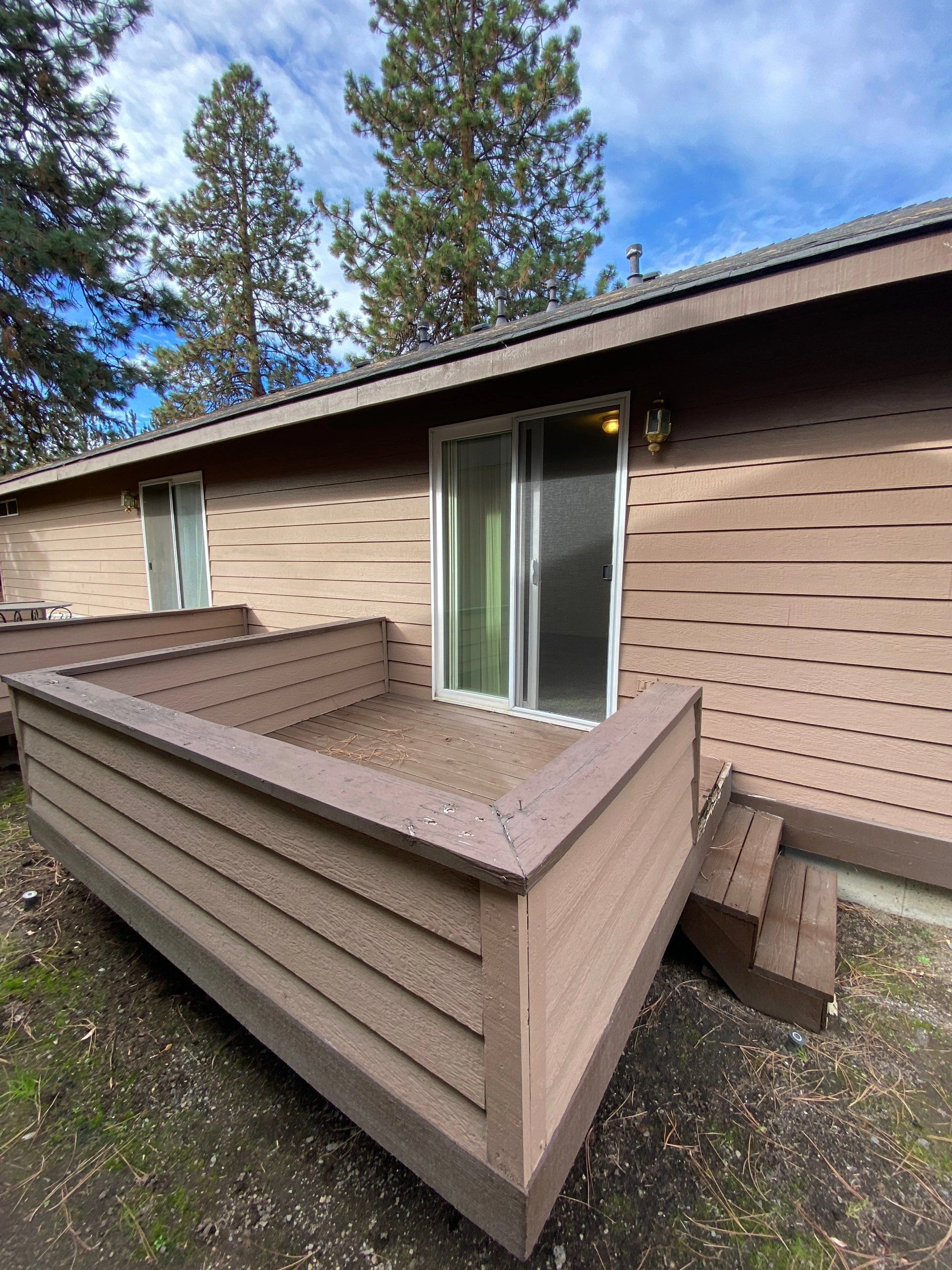 A raised wooden deck with railings sits beside a brown-sided house with sliding glass doors; pine trees and blue sky are visible above.