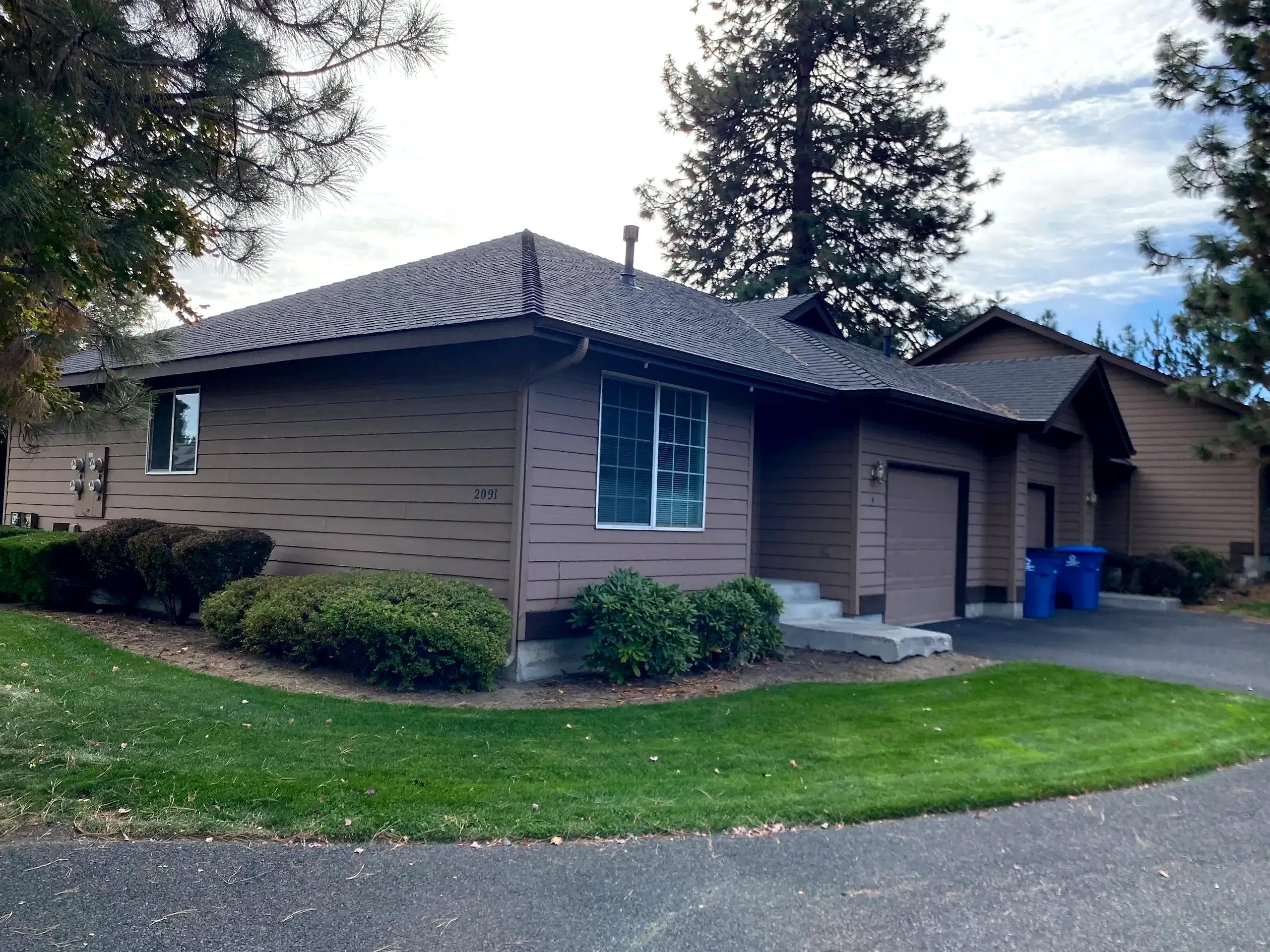A brown-sided building with a dark pitched roof, windows, garage doors, shrubs, and tall trees behind it.