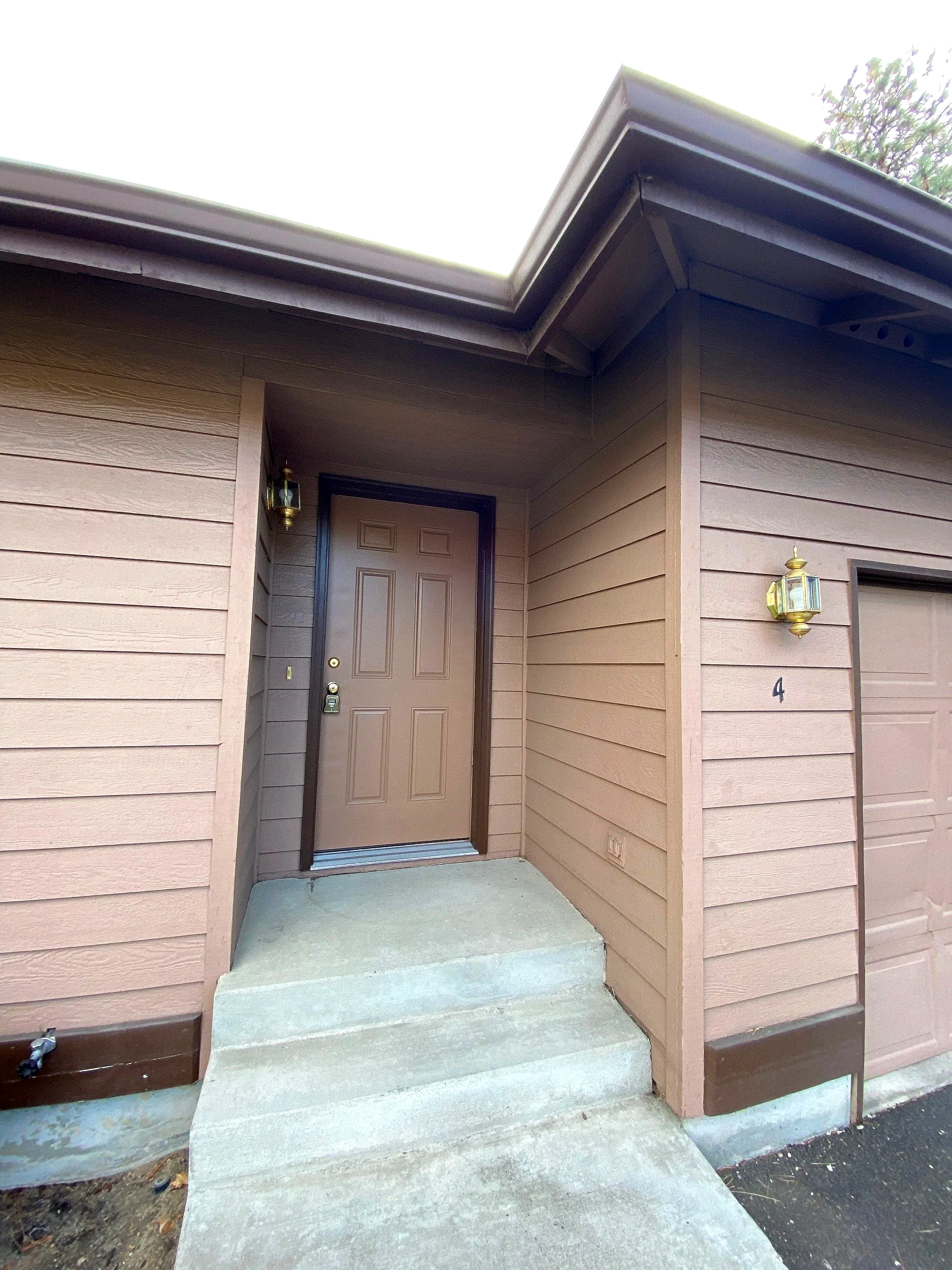 A recessed brown front door beneath a covered porch, with concrete steps, tan siding, and two wall lanterns.
