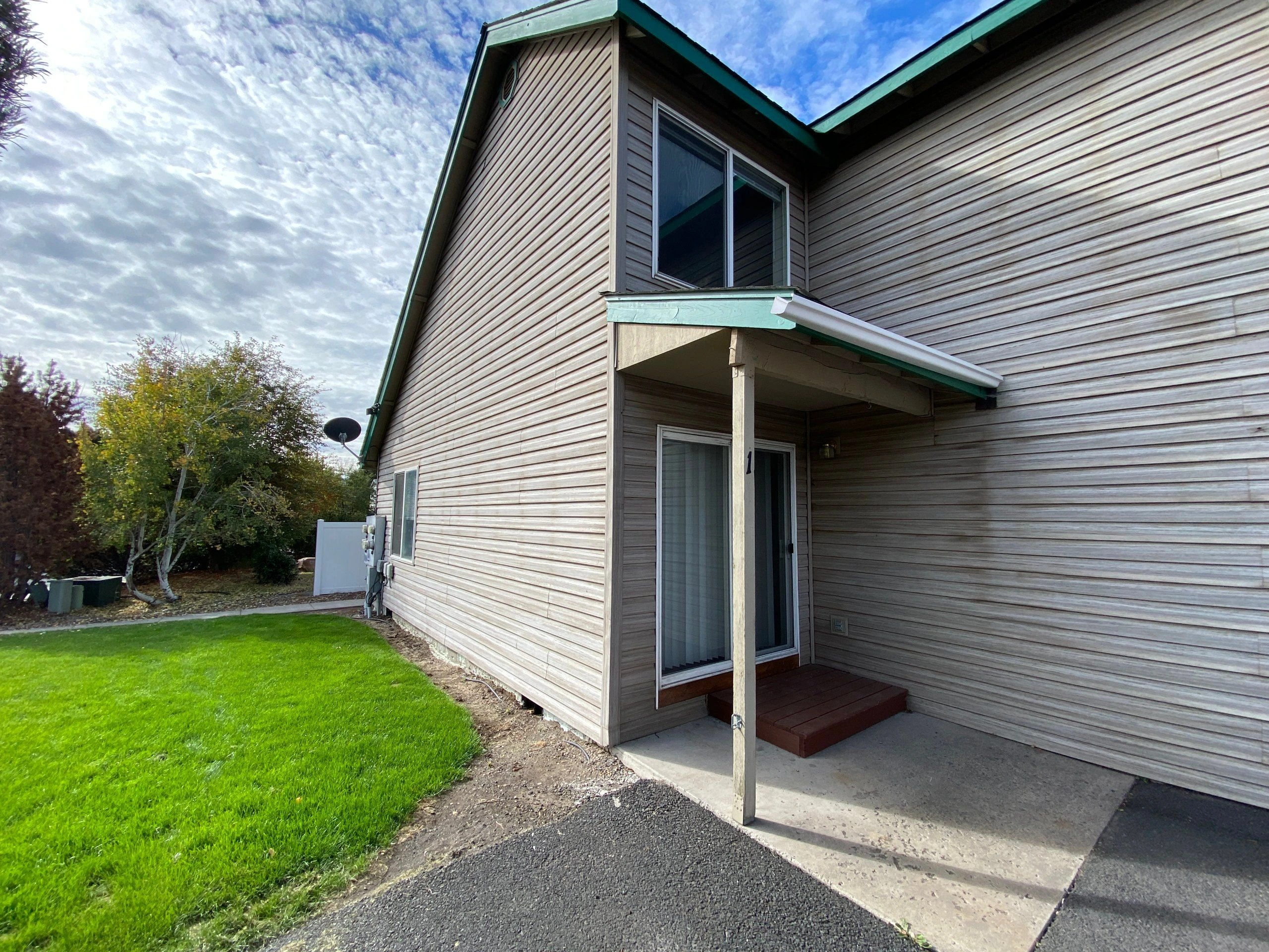 A sided house has a small covered entrance with a post, concrete pad, and sliding glass door beside a green lawn.