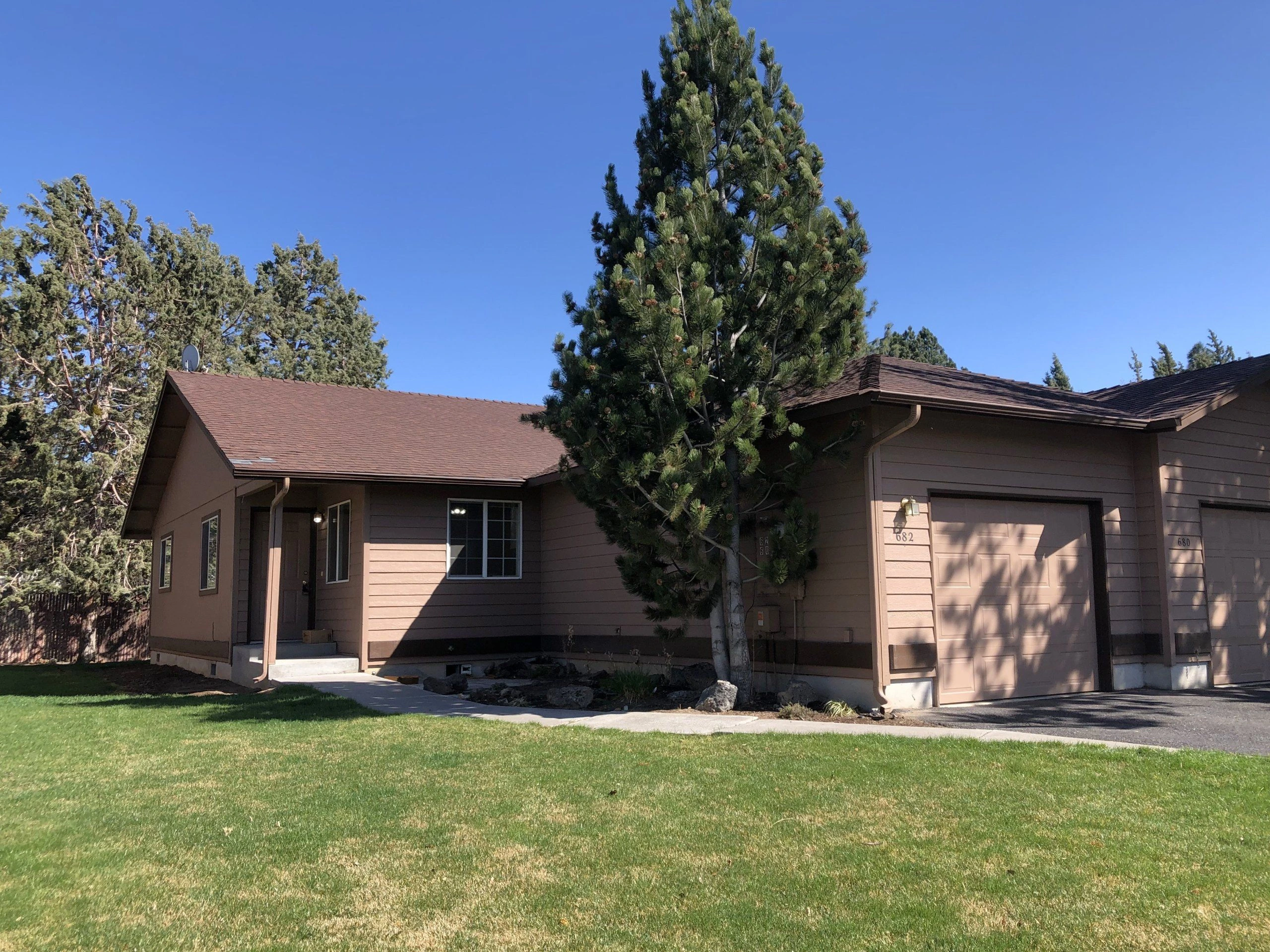 A brown, single-story building with two garage doors stands behind a lawn, partly obscured by a tall pine tree.