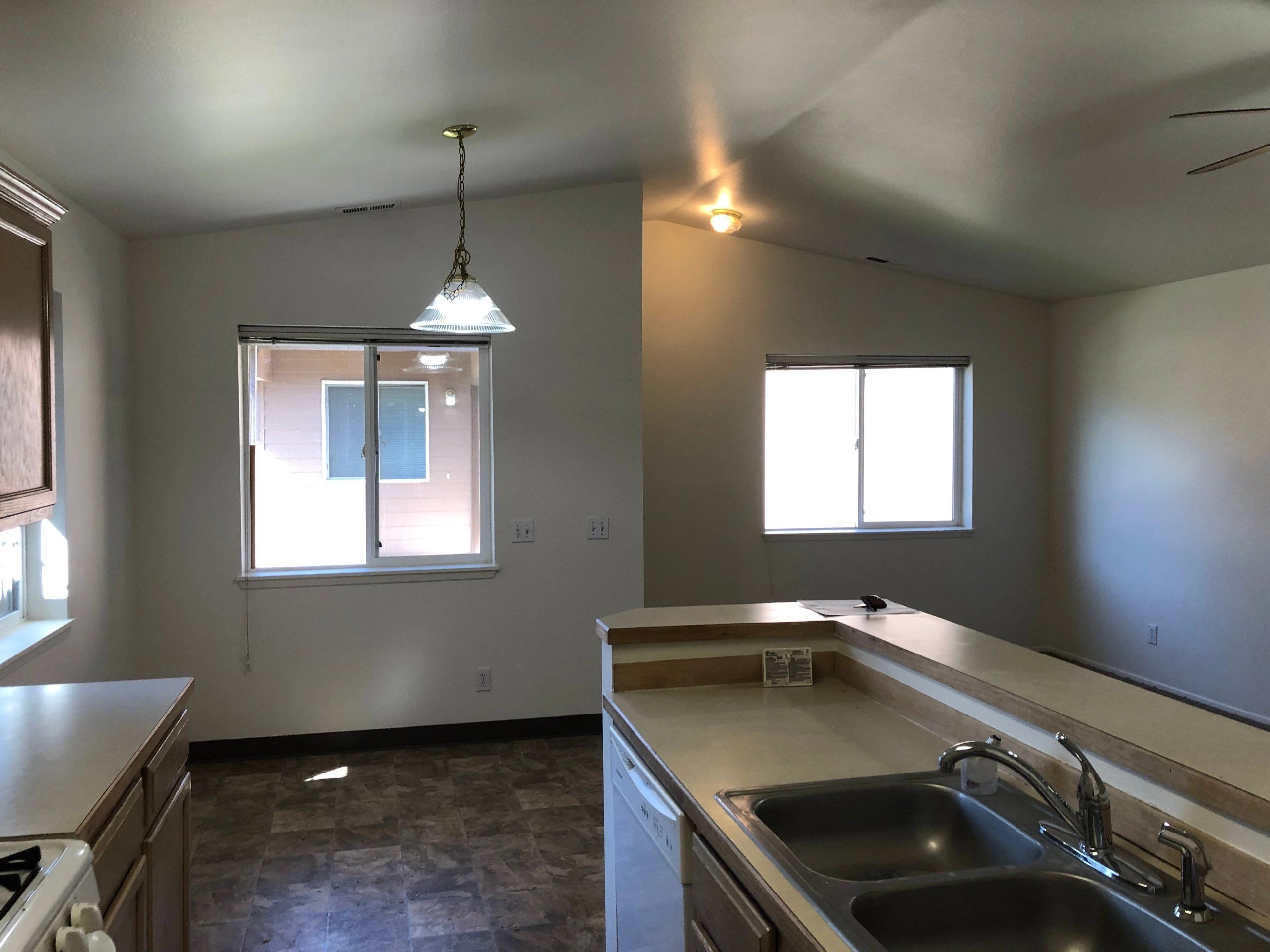 A kitchen with wood cabinets, L-shaped counters and a double sink, with two windows and pendant lighting.