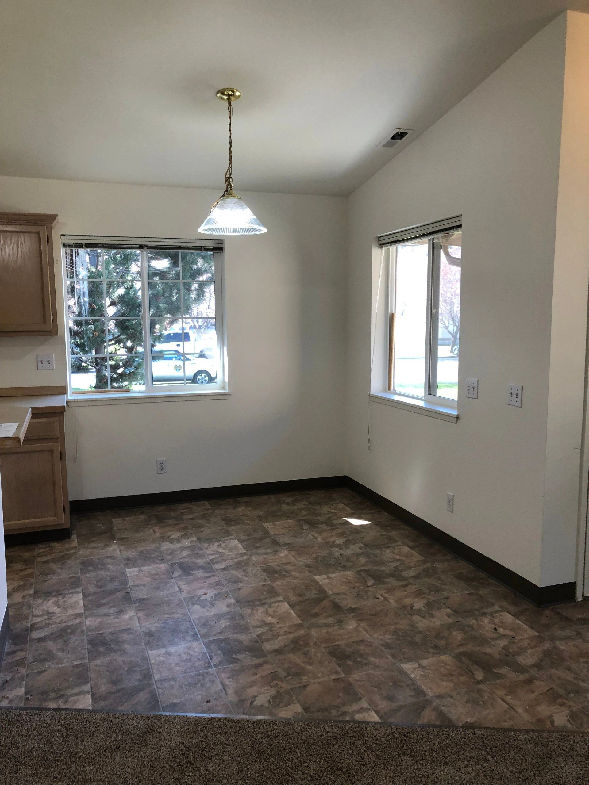 An empty room has tiled flooring, two windows, wooden cabinets, and a hanging ceiling light.