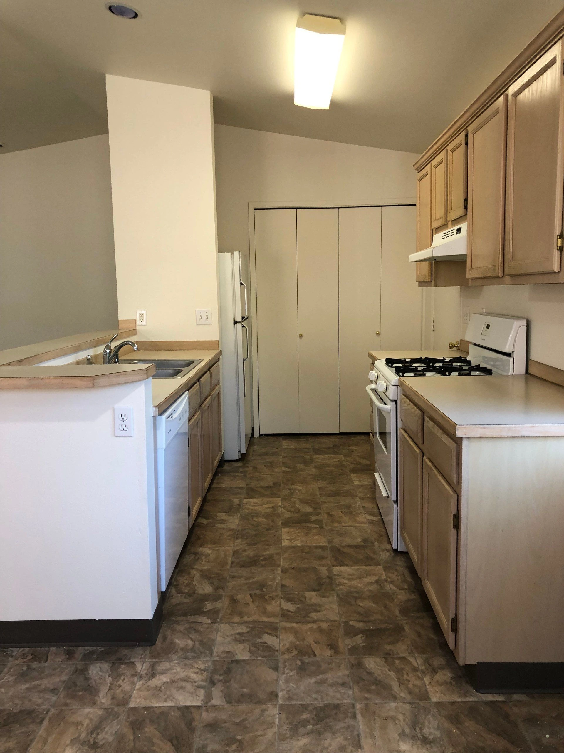 A narrow kitchen has wood cabinets, a double sink, refrigerator, gas stove, and brown patterned tile flooring.