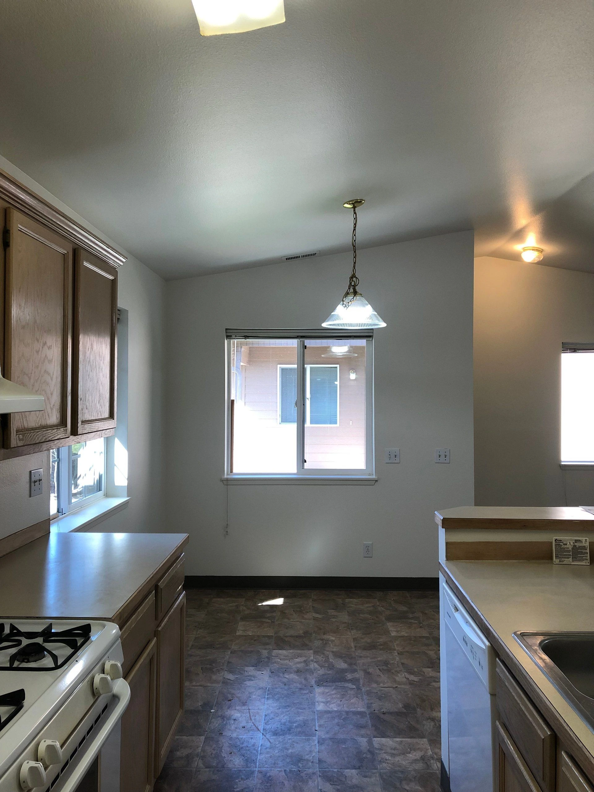 A kitchen with wood cabinets, tiled floor, a stove, counters, a sink, windows, and a hanging ceiling light.
