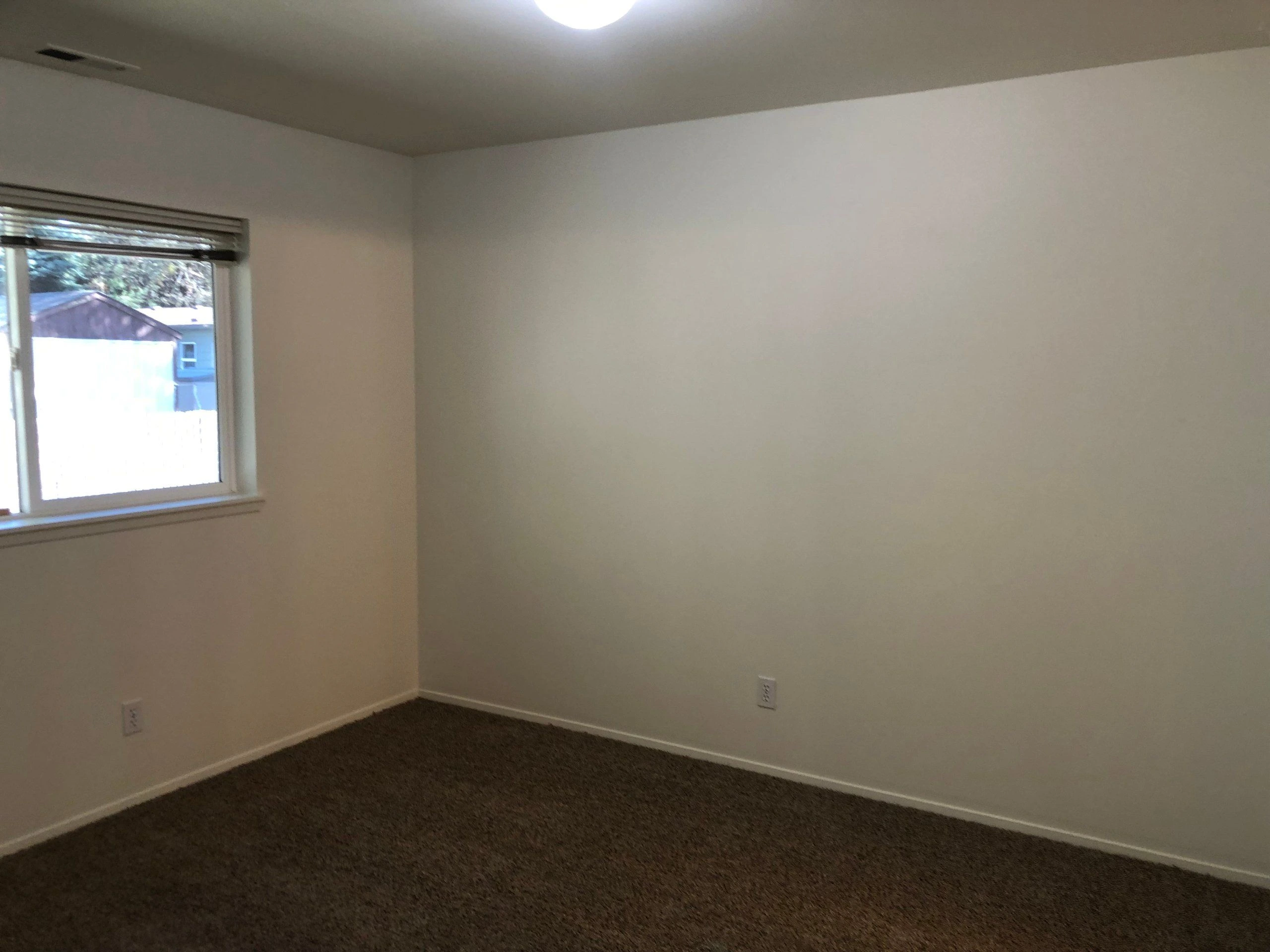 An empty room with beige walls, brown carpet, a window with blinds, and a recessed ceiling.