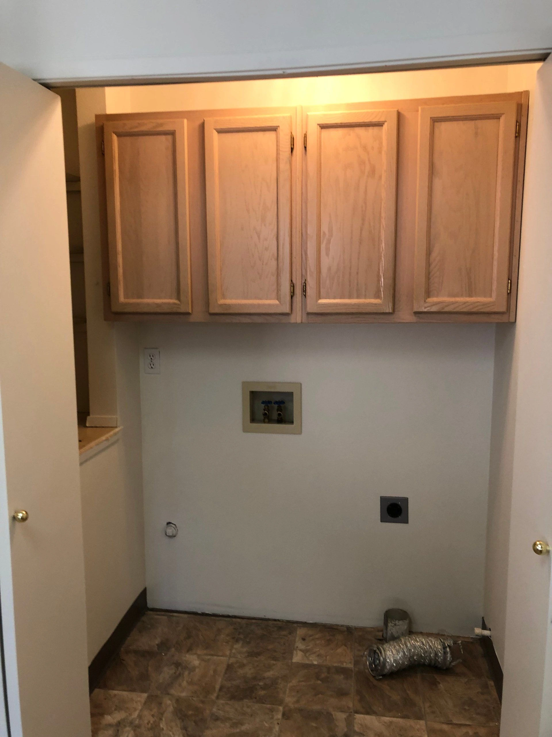 Four wooden cabinet doors hang above a recessed wall with outlets and fittings; a flexible metal duct lies on the tiled floor.