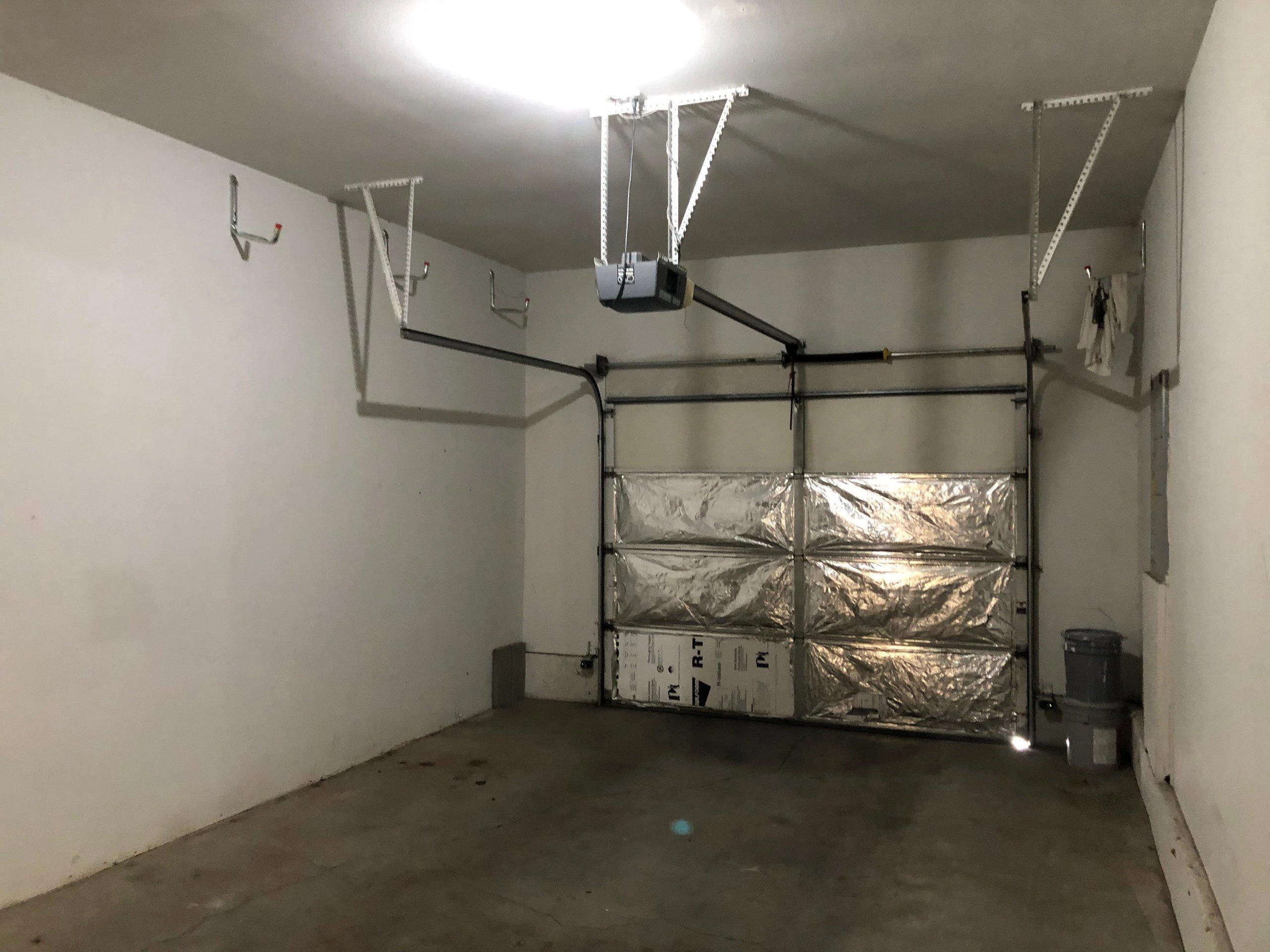 An empty garage with a closed segmented door, ceiling-mounted opener and rails, wall hooks, and a concrete floor.