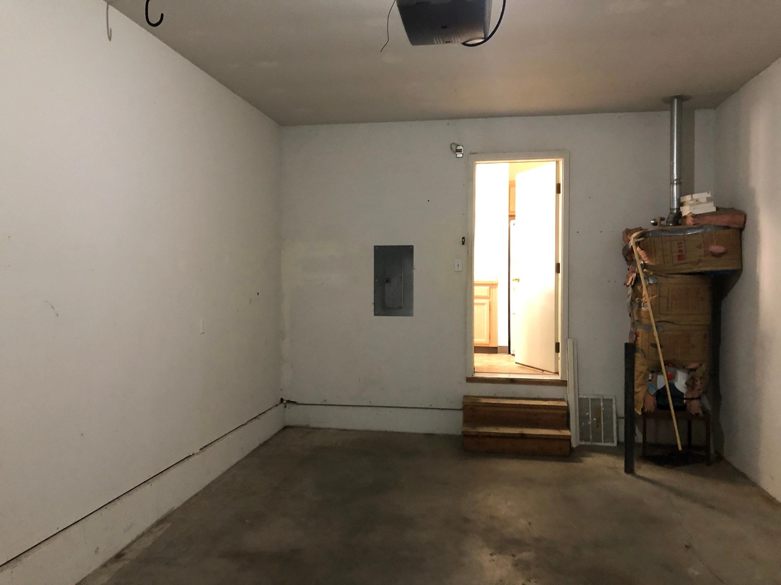 A bare room with a concrete floor, an open doorway reached by two wooden steps, and stacked items beside a metal pipe.