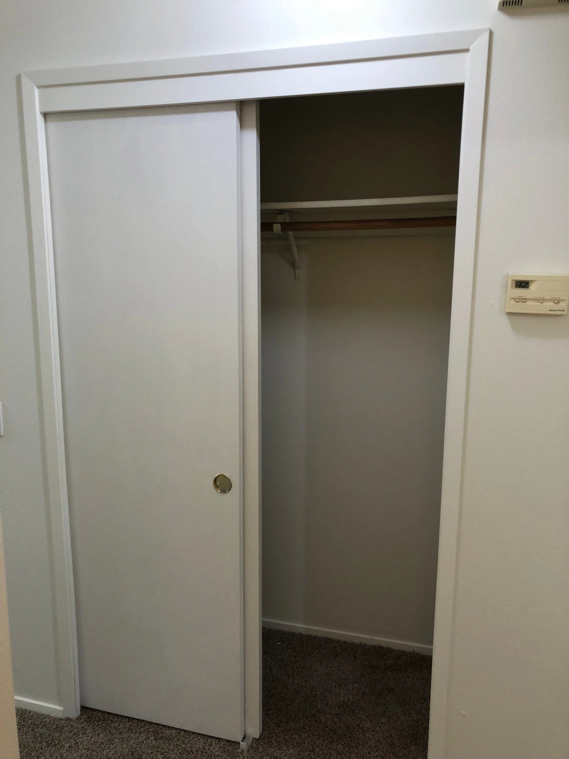 An open closet has a white sliding door, a wooden hanging rod, and carpeted flooring.