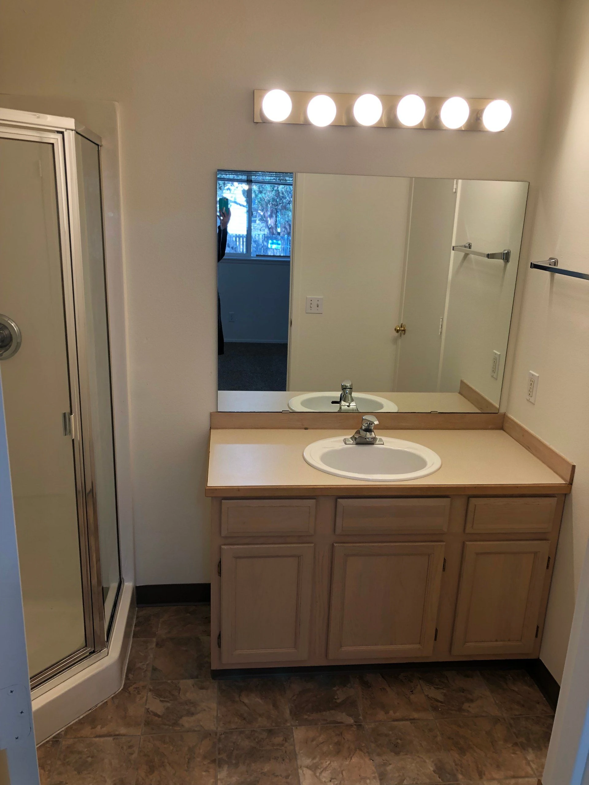 A bathroom vanity with a white sink, wood cabinets, a large mirror, six lights, and a glass shower enclosure.