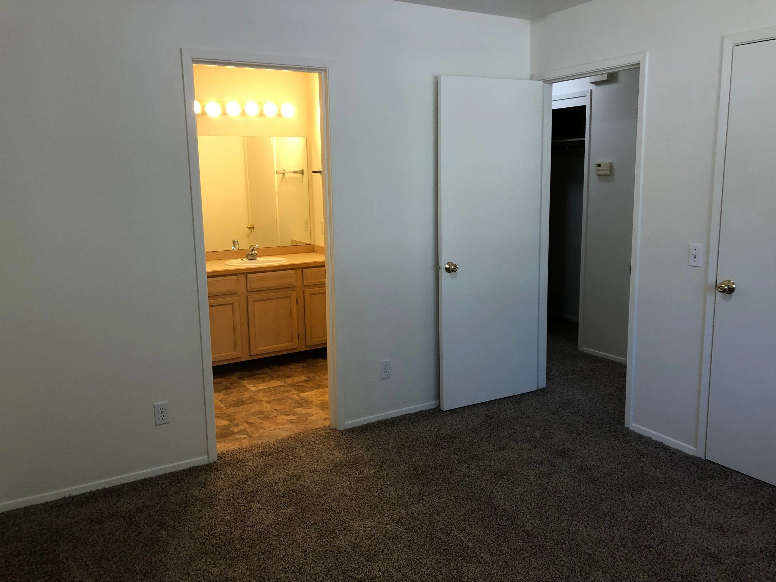 A carpeted room has open doors to a brightly lit vanity area and a darker adjoining space.