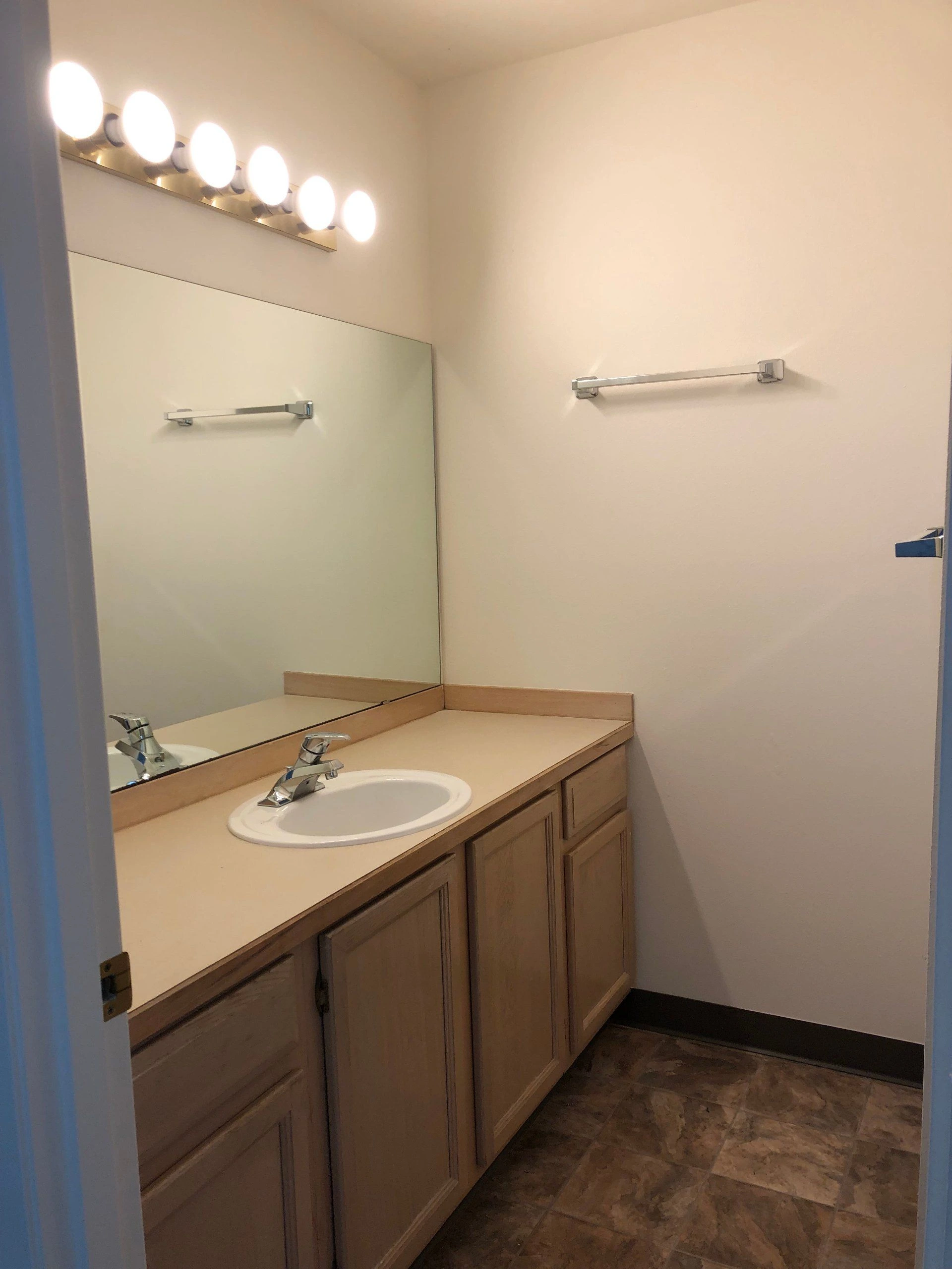 A bathroom vanity has a white oval sink, wood cabinets, a large mirror, and a row of bright lights above.