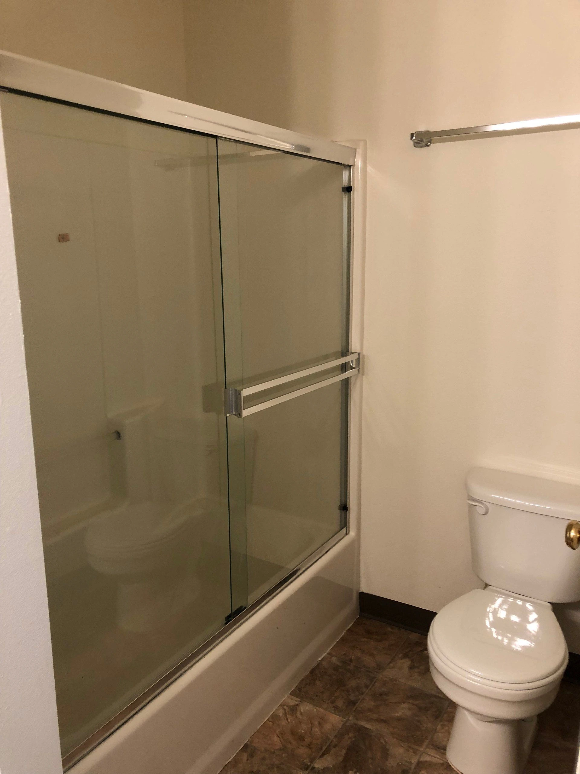 A glass shower enclosure with metal framing stands beside a white toilet on a brown tiled floor.