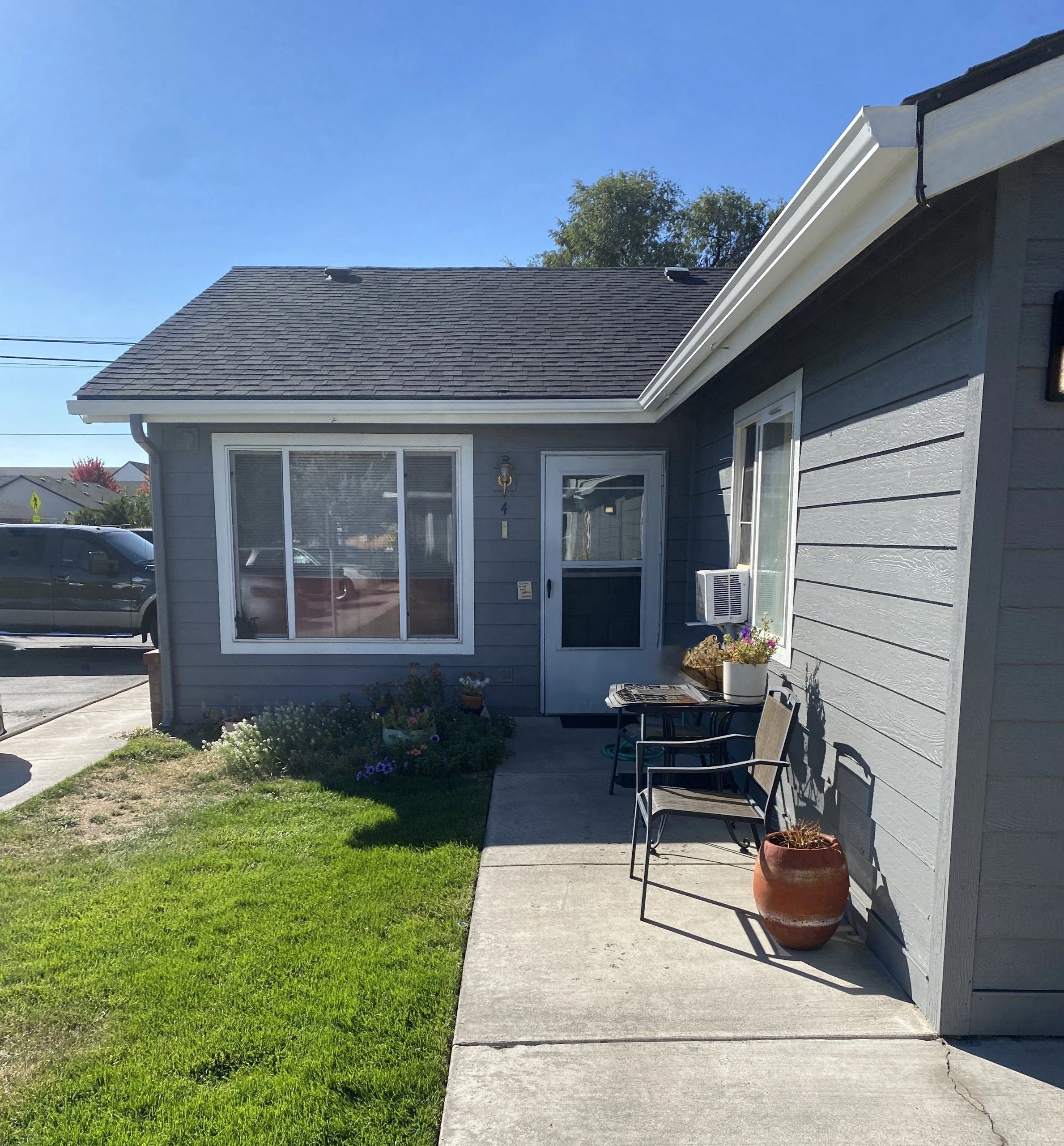 A gray-sided house with a shingled roof, white-trimmed windows, and a door faces a concrete patio with chairs and potted plants.
