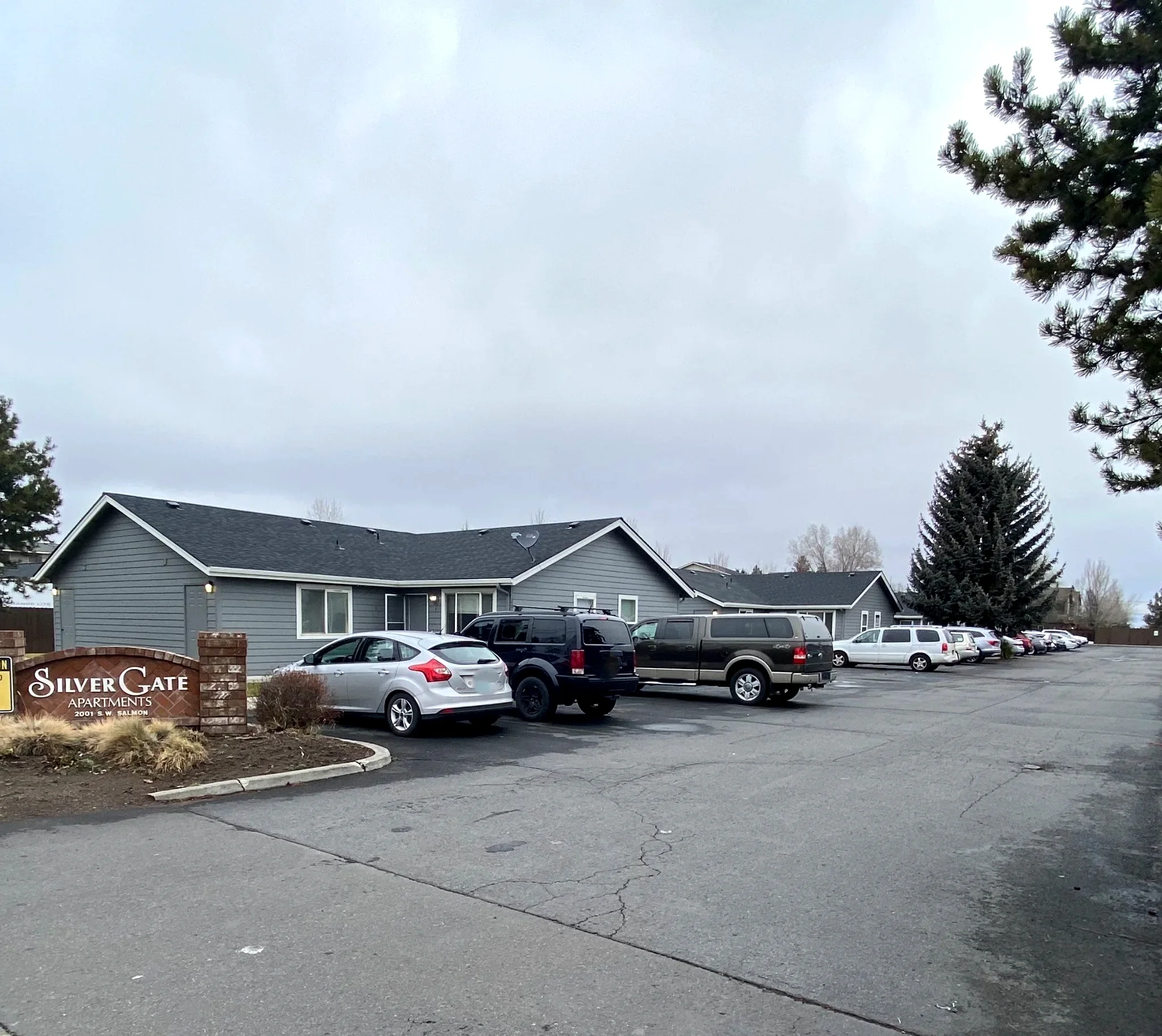 A gray, single-story building stands beside a parking lot with several cars and a sign reading “Silver Gate Apartments.”