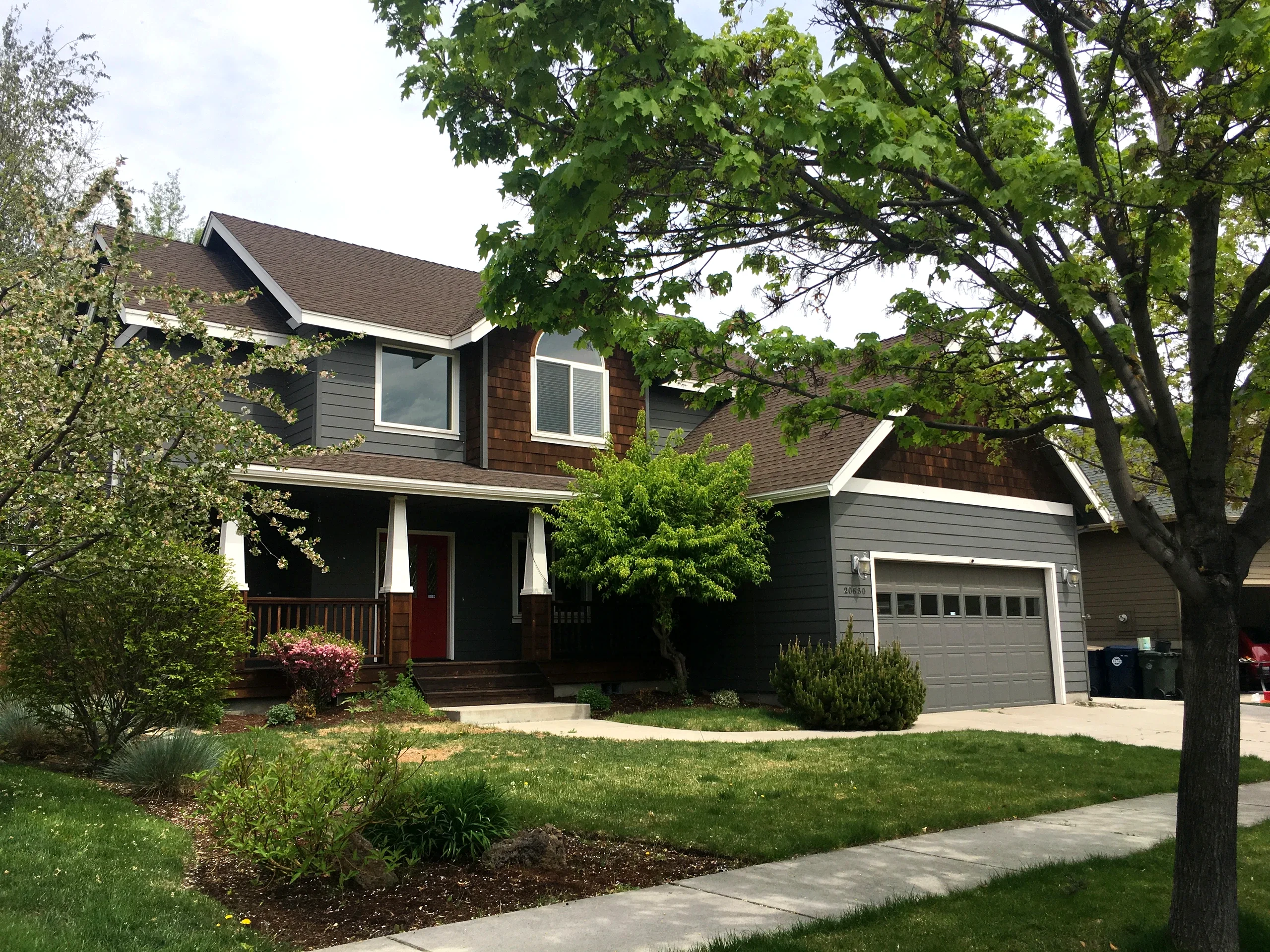A two-story house with gray siding, brown shingle accents, a red front door, and an attached garage stands behind leafy trees and a lawn.