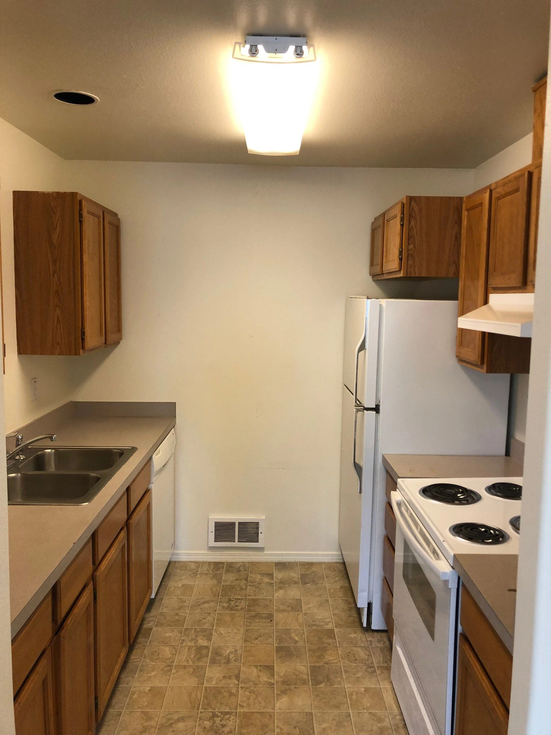 A narrow kitchen has wooden cabinets, a double sink, a refrigerator, an electric stove, and a tiled floor.