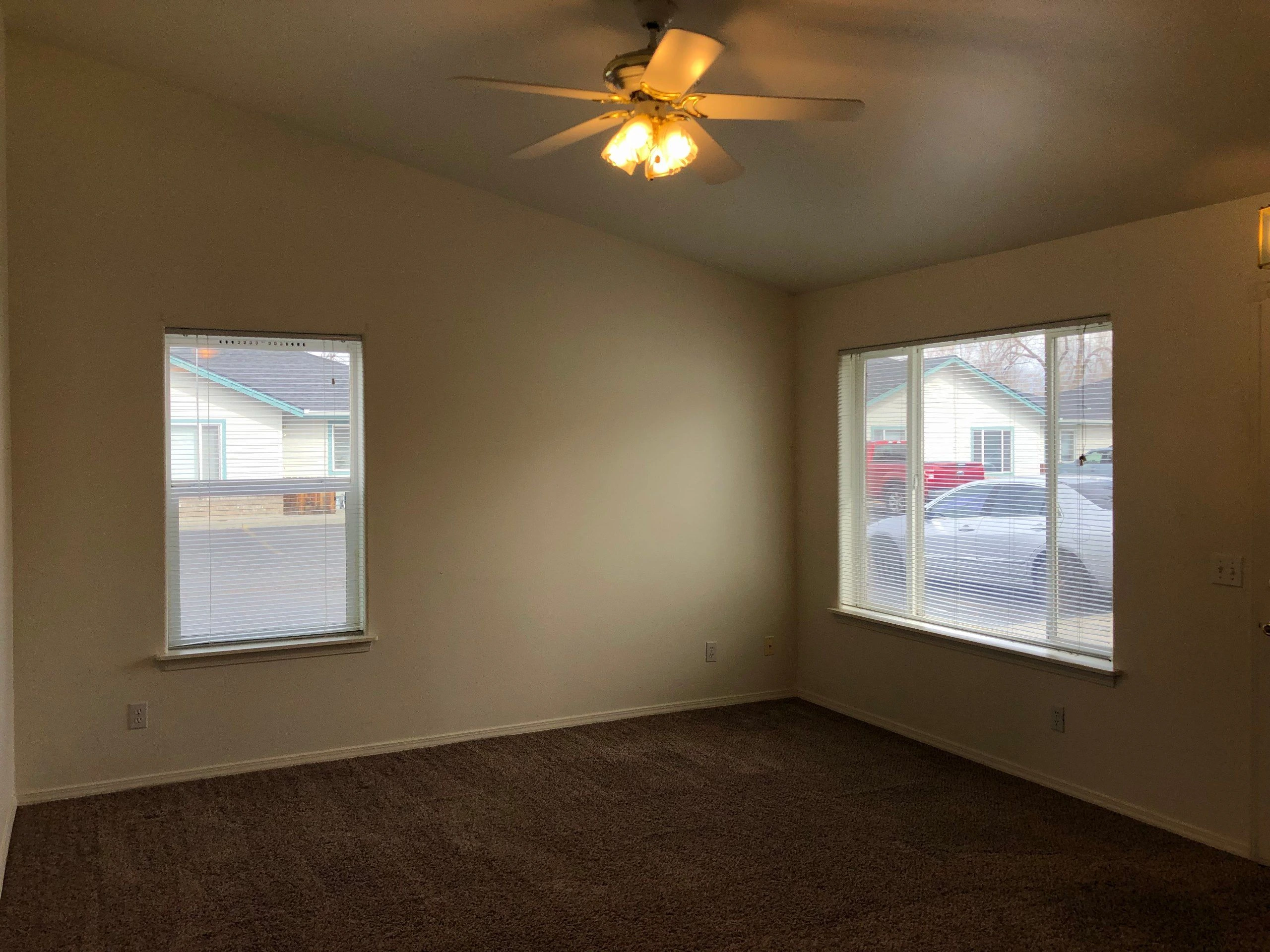 An empty room with beige walls, brown carpet, two windows with blinds, and a lit ceiling fan.