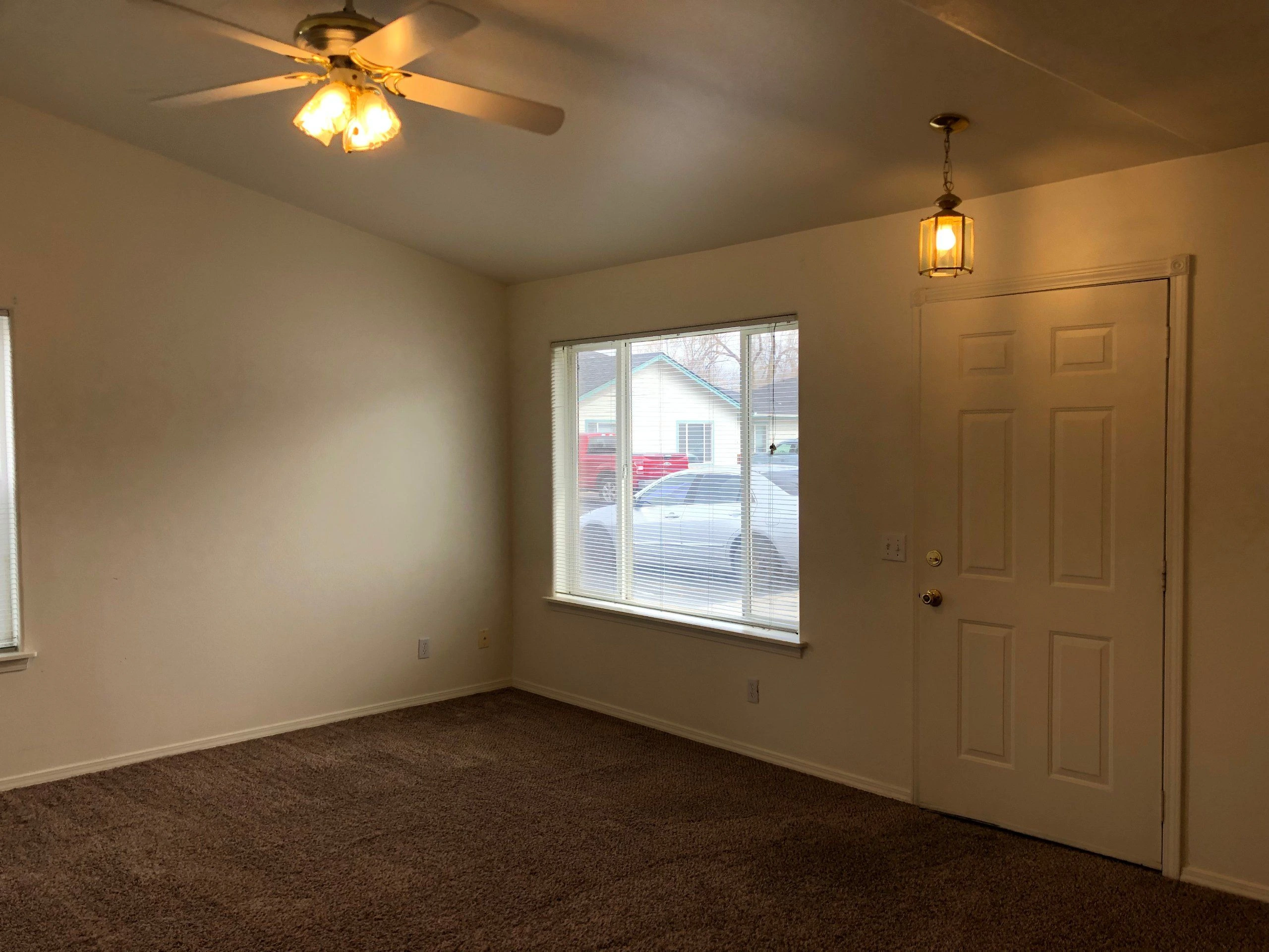 An empty room with brown carpet, light walls, a window with blinds, a paneled door, and two ceiling lights.