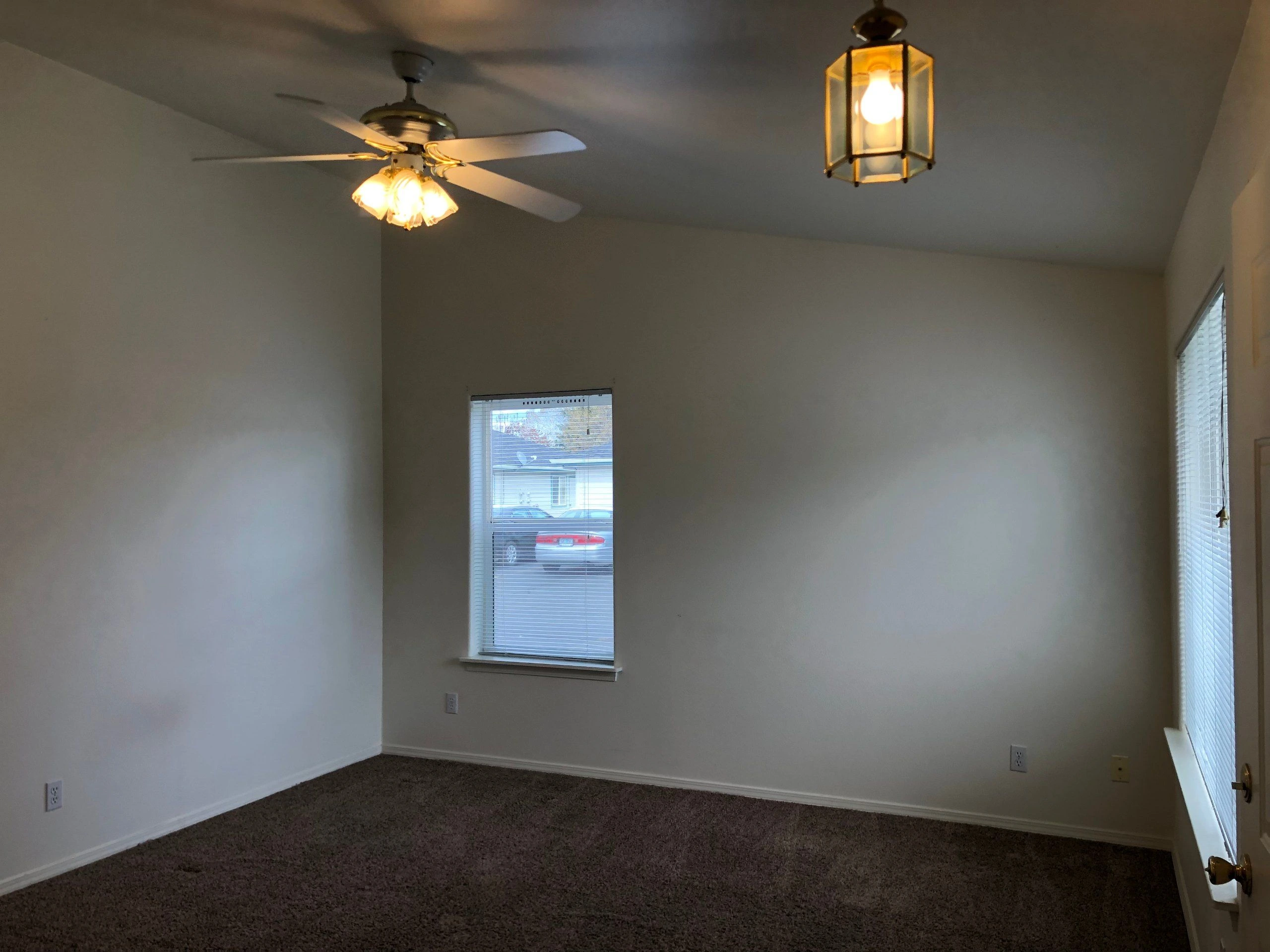 An empty room with carpet, pale walls, two windows with blinds, a ceiling fan with lights, and a hanging lantern.