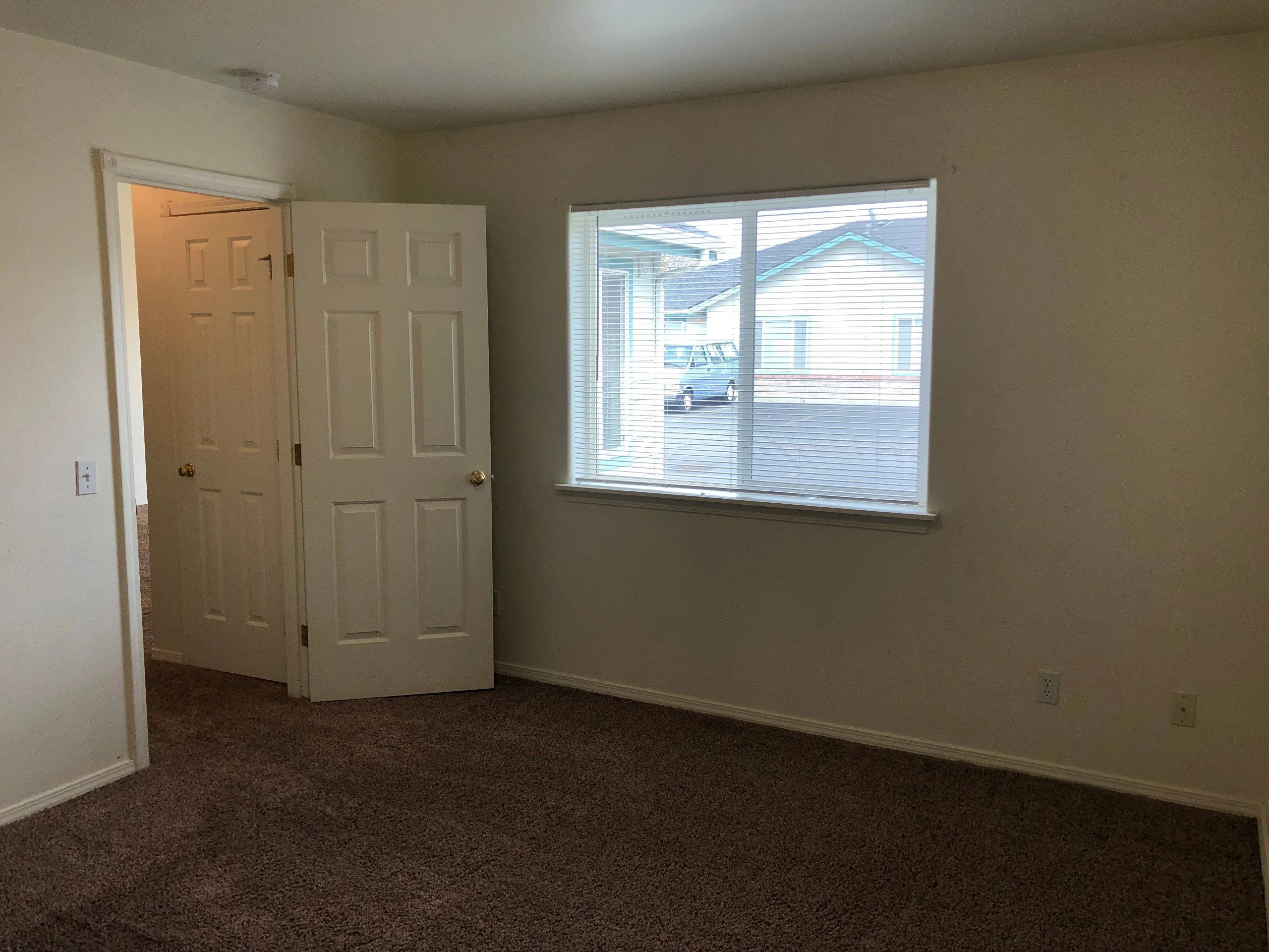 An empty room with brown carpet, light walls, an open white door, and a window with horizontal blinds.